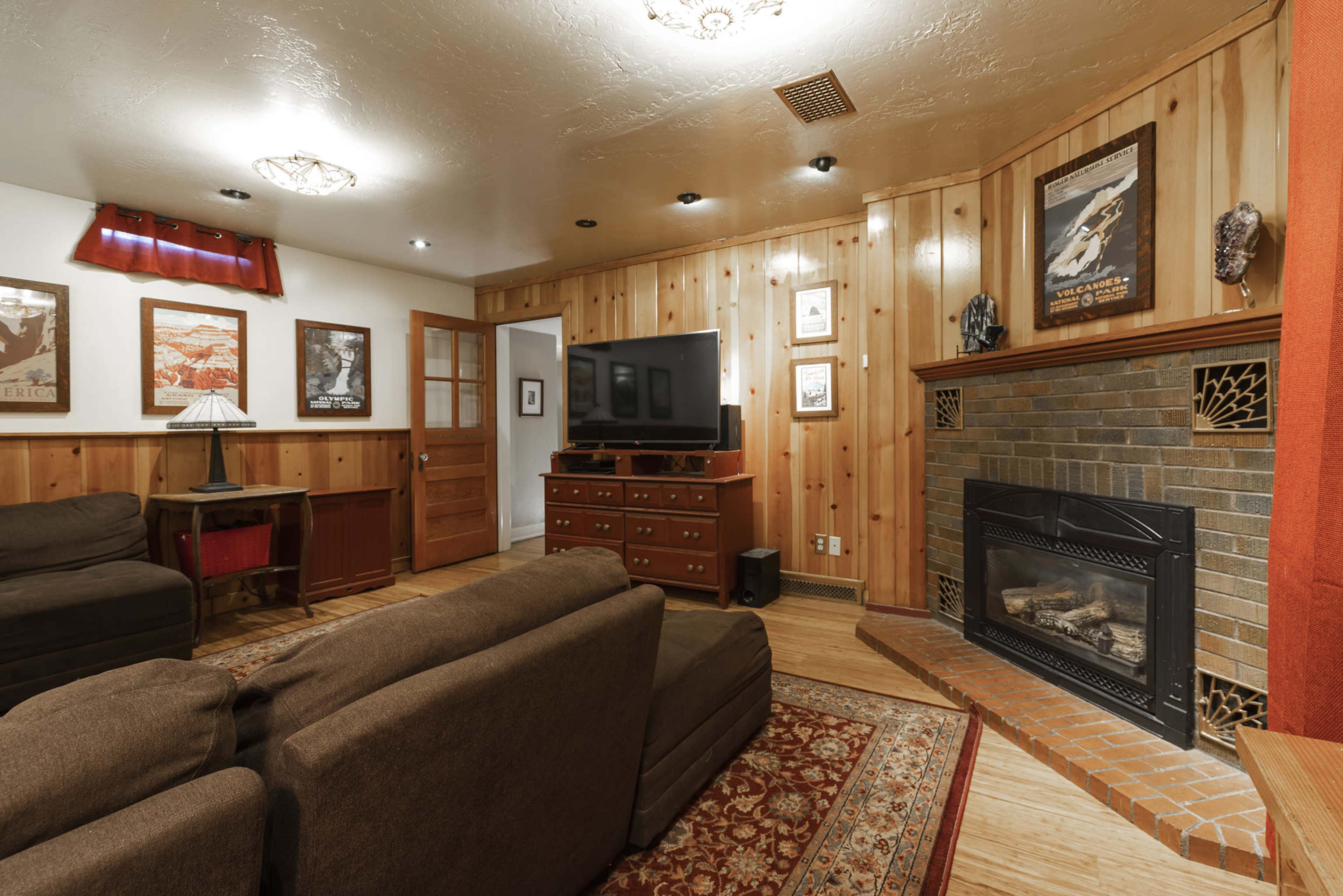 The room features wooden paneling, a brick fireplace, a television on a wooden cabinet, and two brown sofas arranged around a patterned area rug.