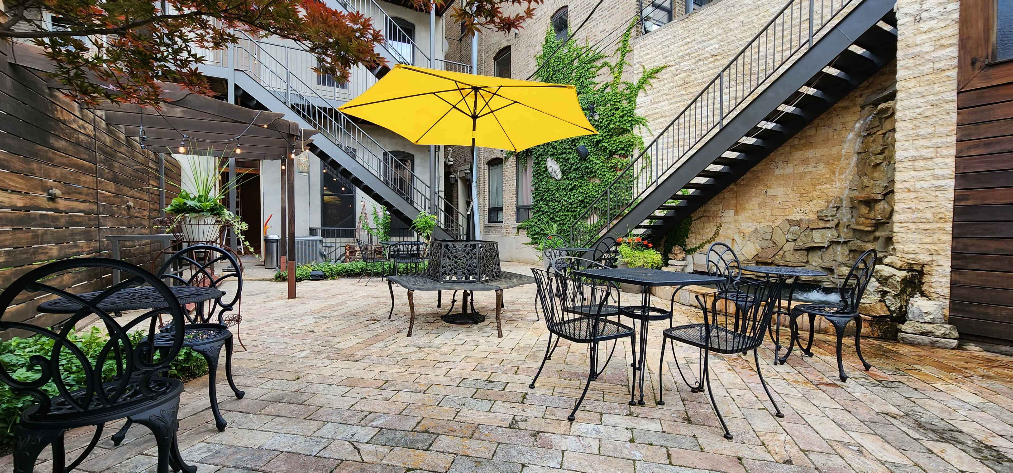 A stone-paved courtyard features black metal tables and chairs under a yellow umbrella, surrounded by greenery and brick walls.