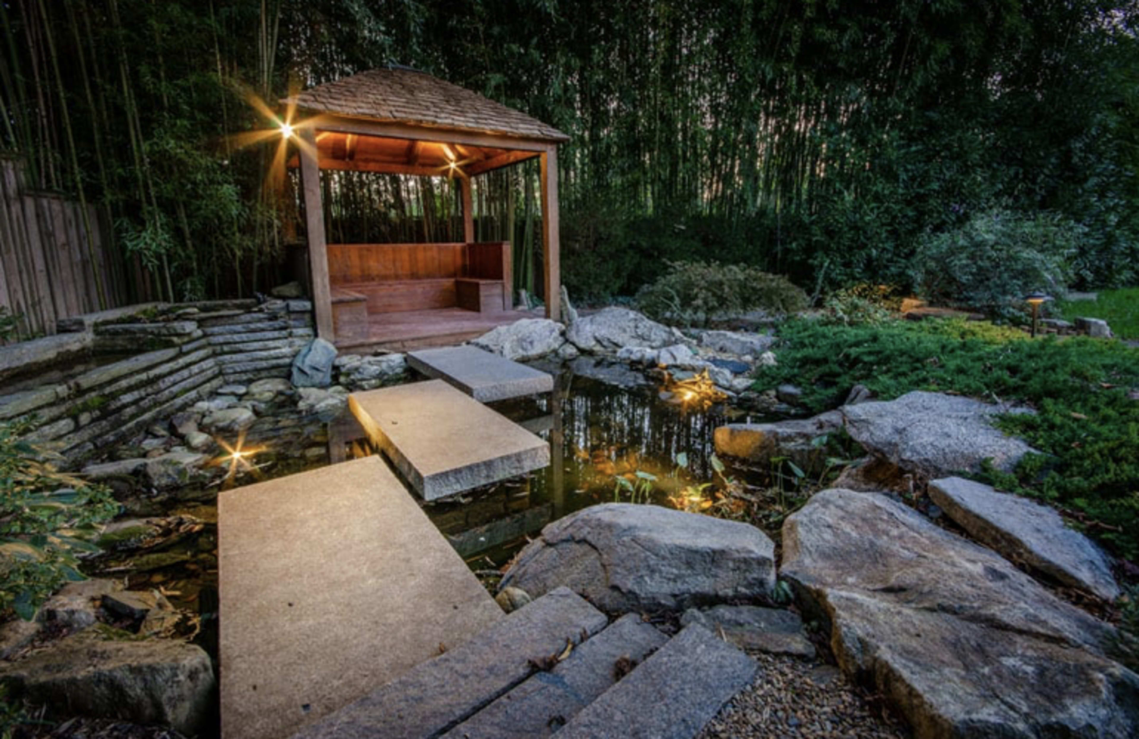 The image depicts a serene garden scene featuring a wooden gazebo surrounded by rocks, a pond, and stone pathways illuminated by soft lights.
