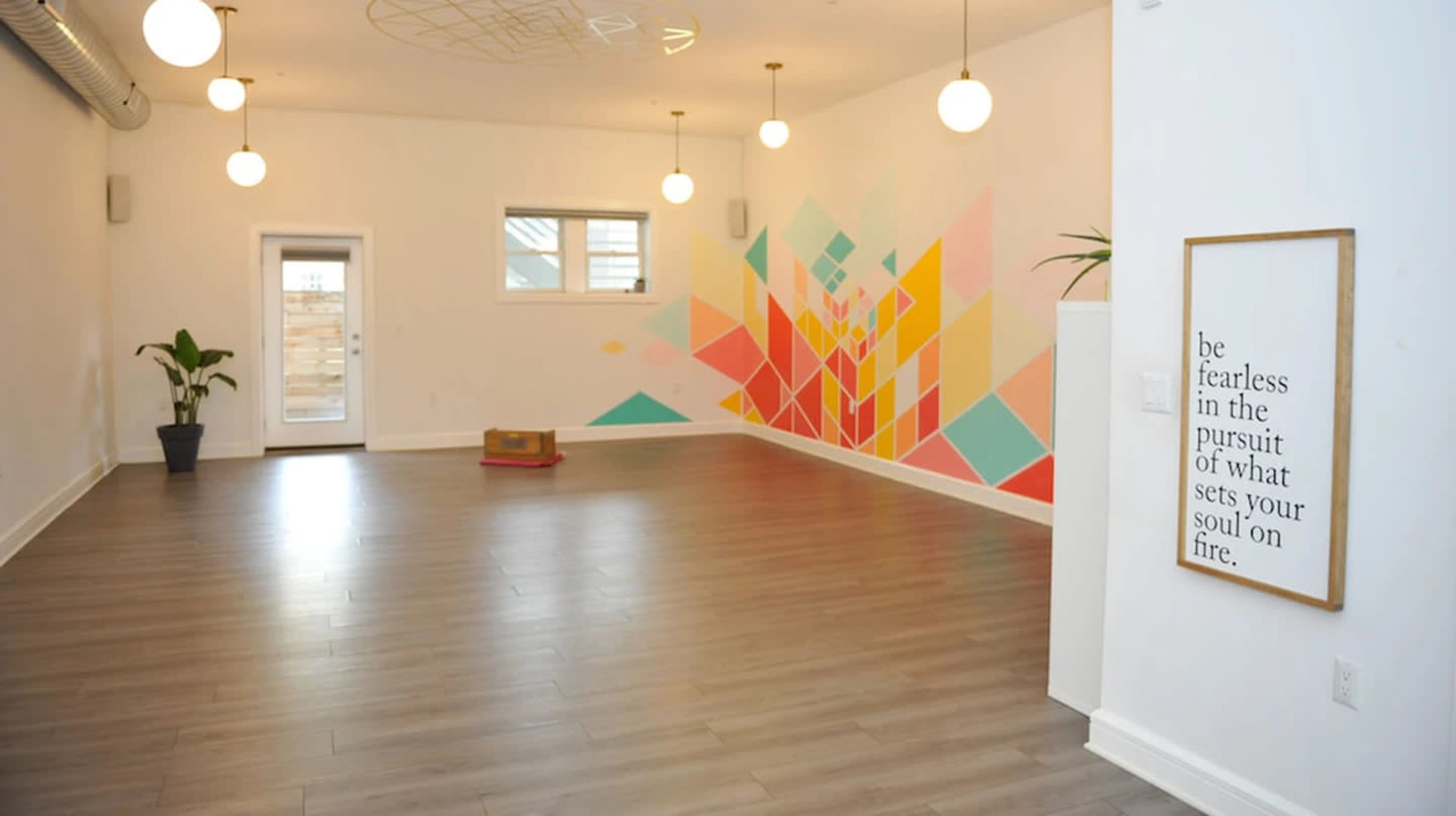 The room features a light wooden floor, a colorful geometric mural on the wall, and pendant lights hanging from the ceiling.