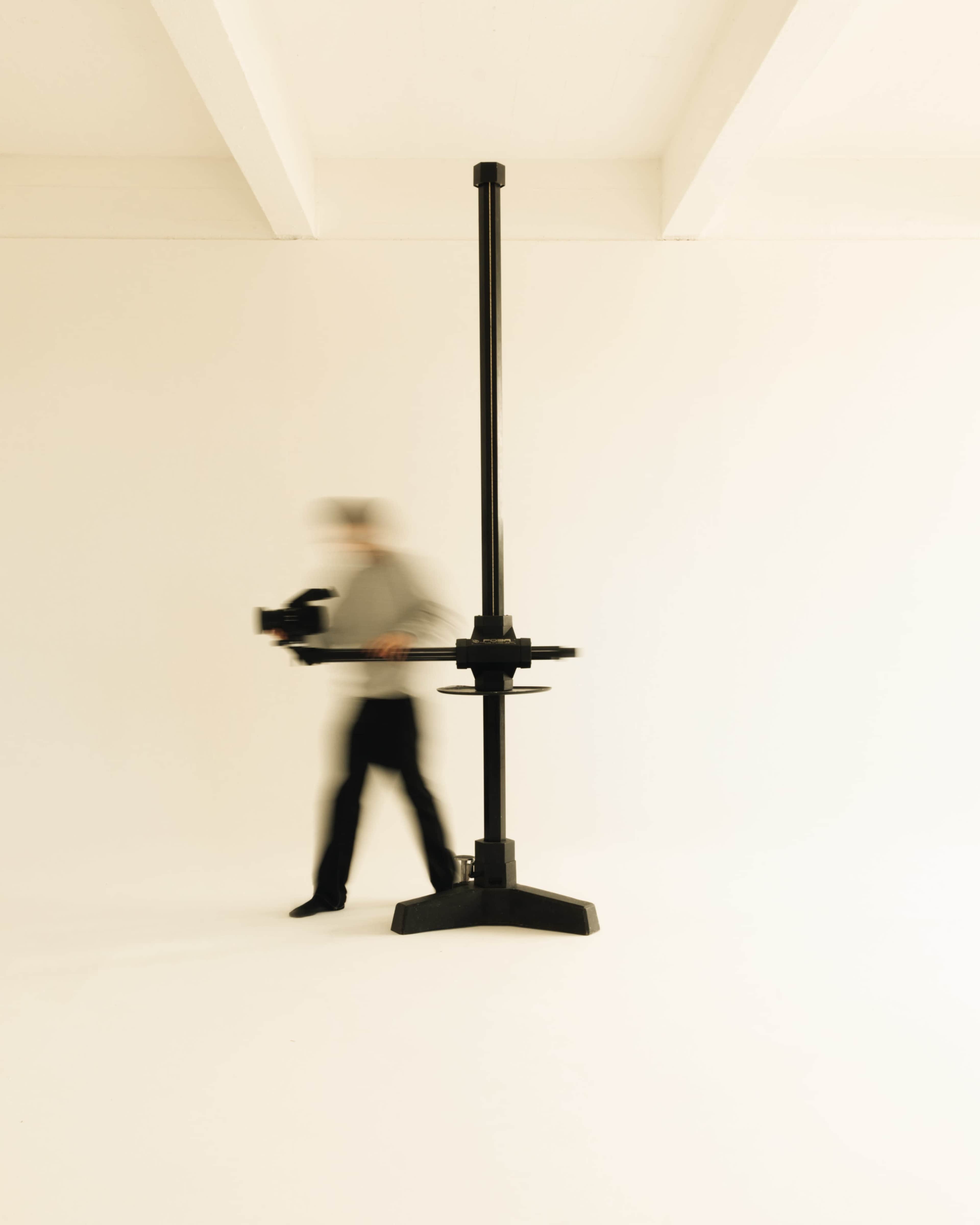 A person walks past a tall, black studio stand in a minimalistic, white room.