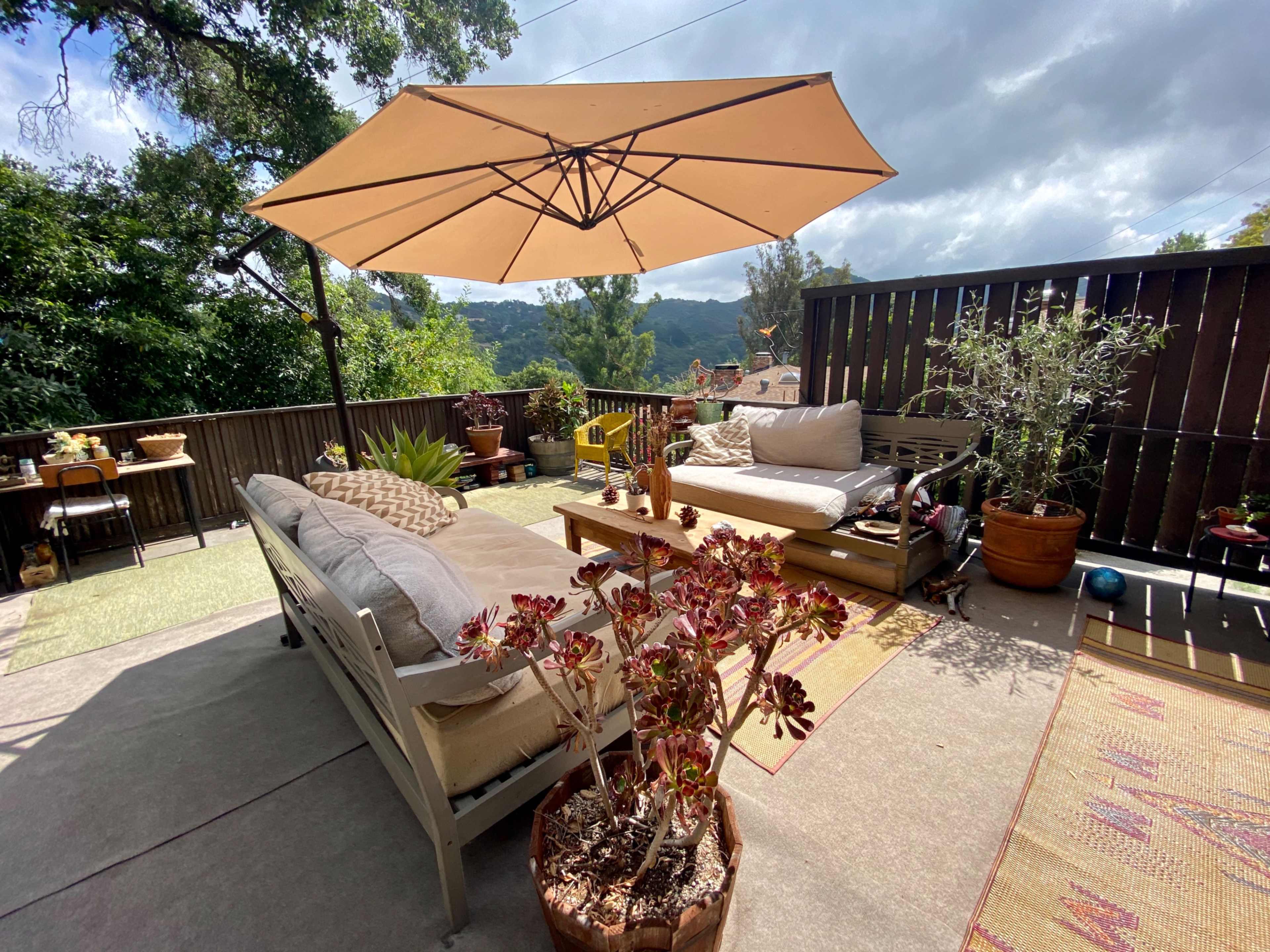 A spacious outdoor patio features a shaded seating area with a beige umbrella, surrounded by various plants and decorative pots.