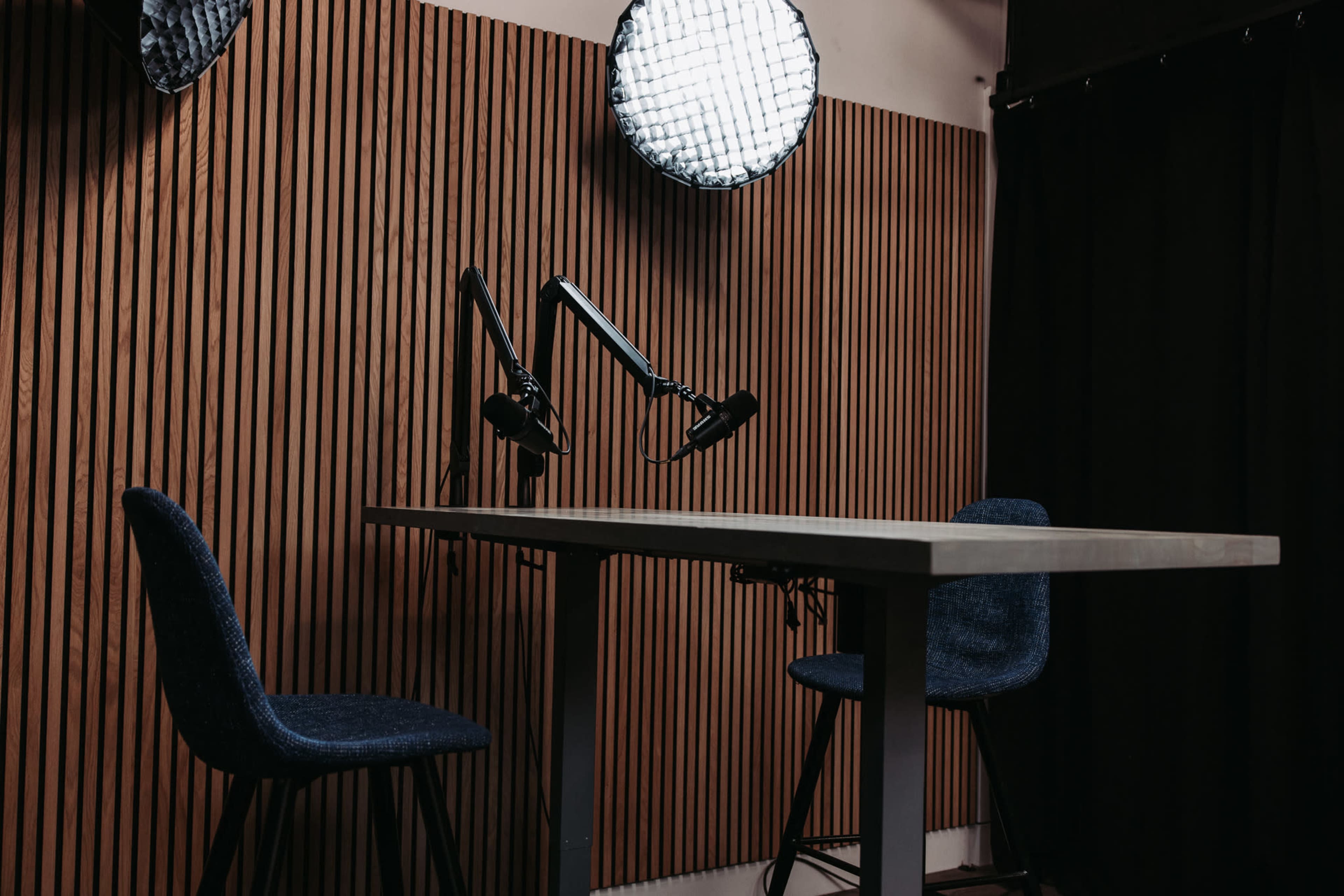 A minimalistic recording studio features a table with two chairs and mounted microphones, set against a wooden panel backdrop.