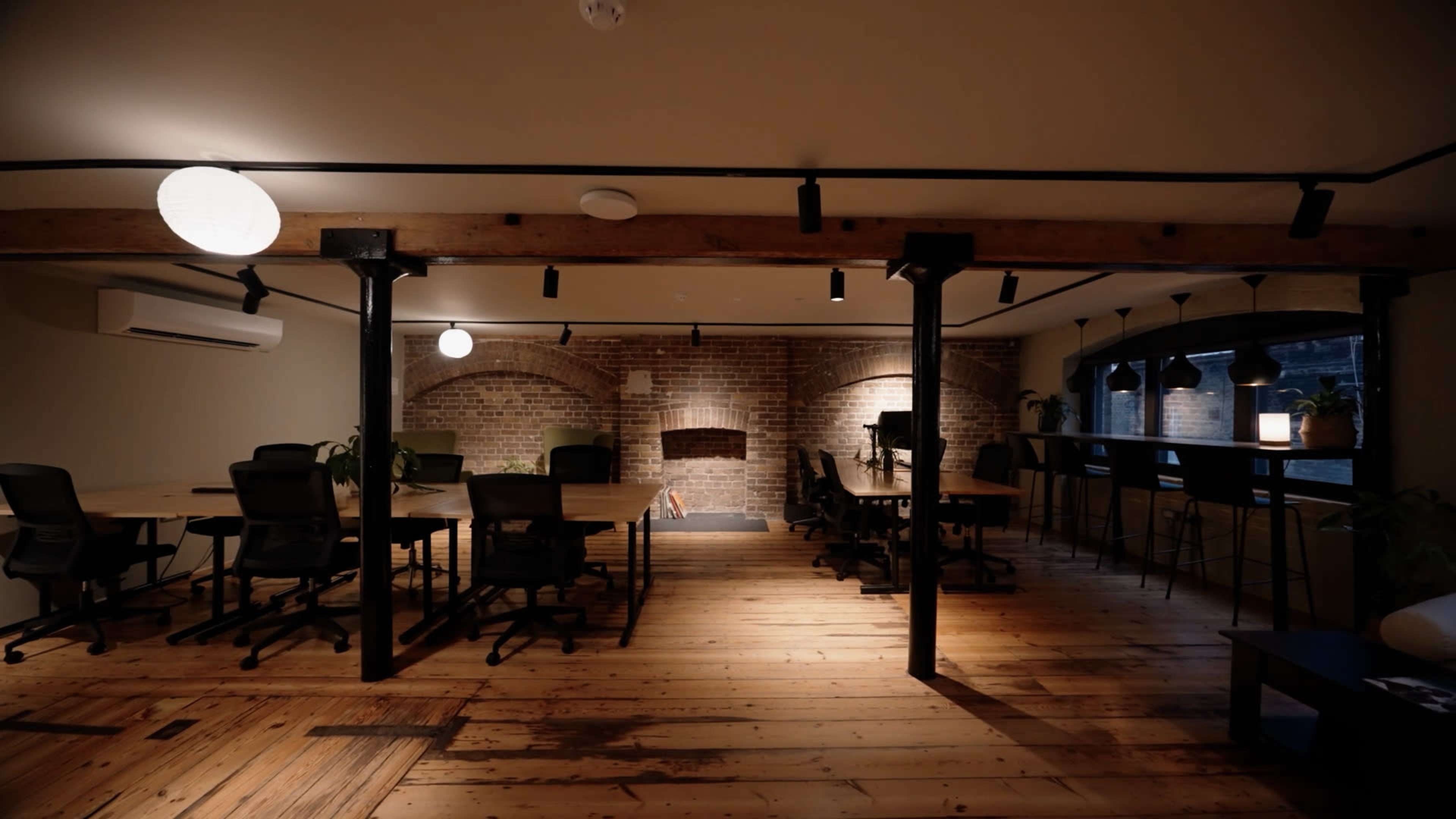 The image shows a spacious modern office with wooden floors, several desks and chairs, and a brick wall with a fireplace.