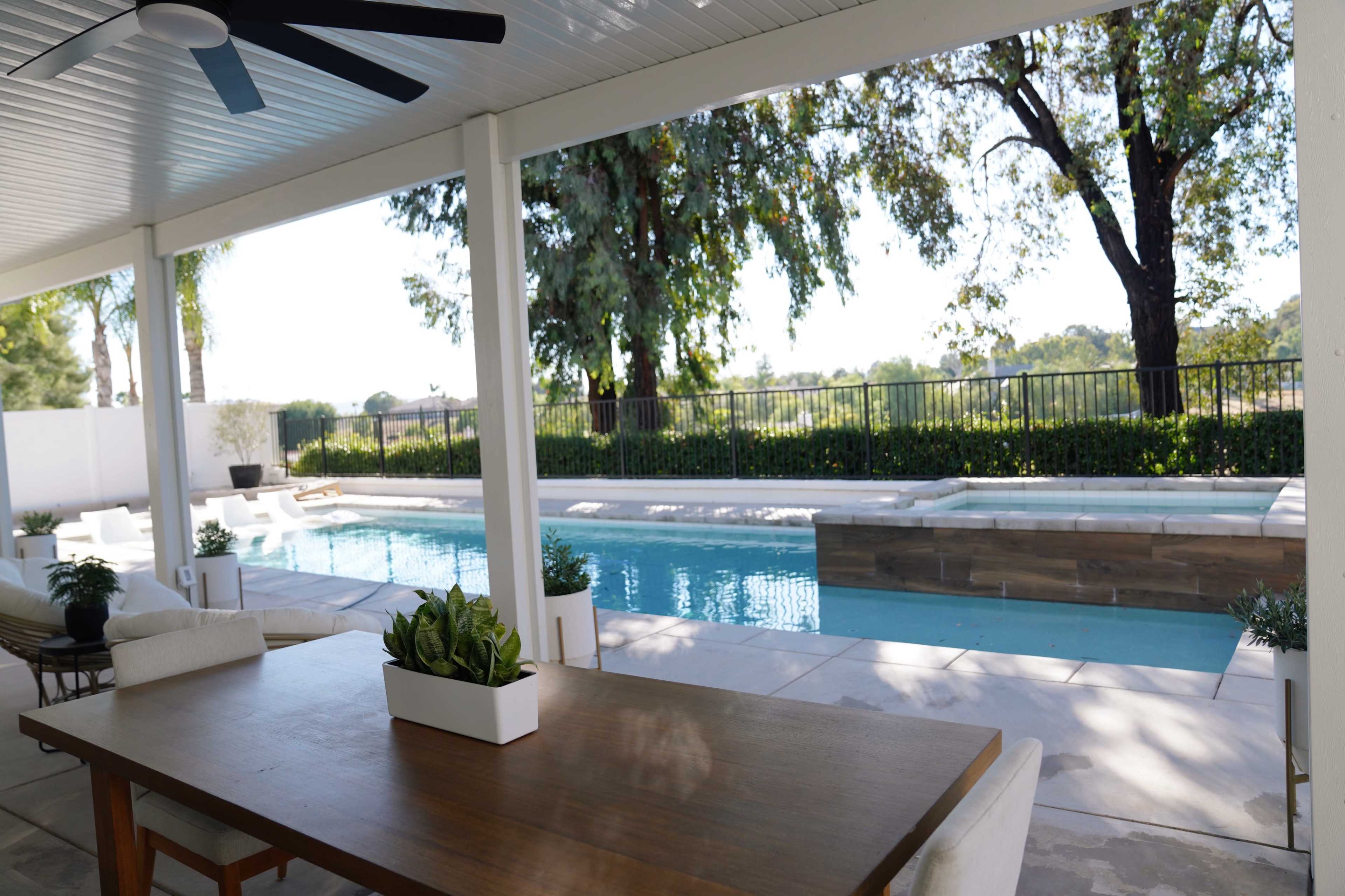 A spacious patio features a dining table overlooking a swimming pool surrounded by greenery and a fence.