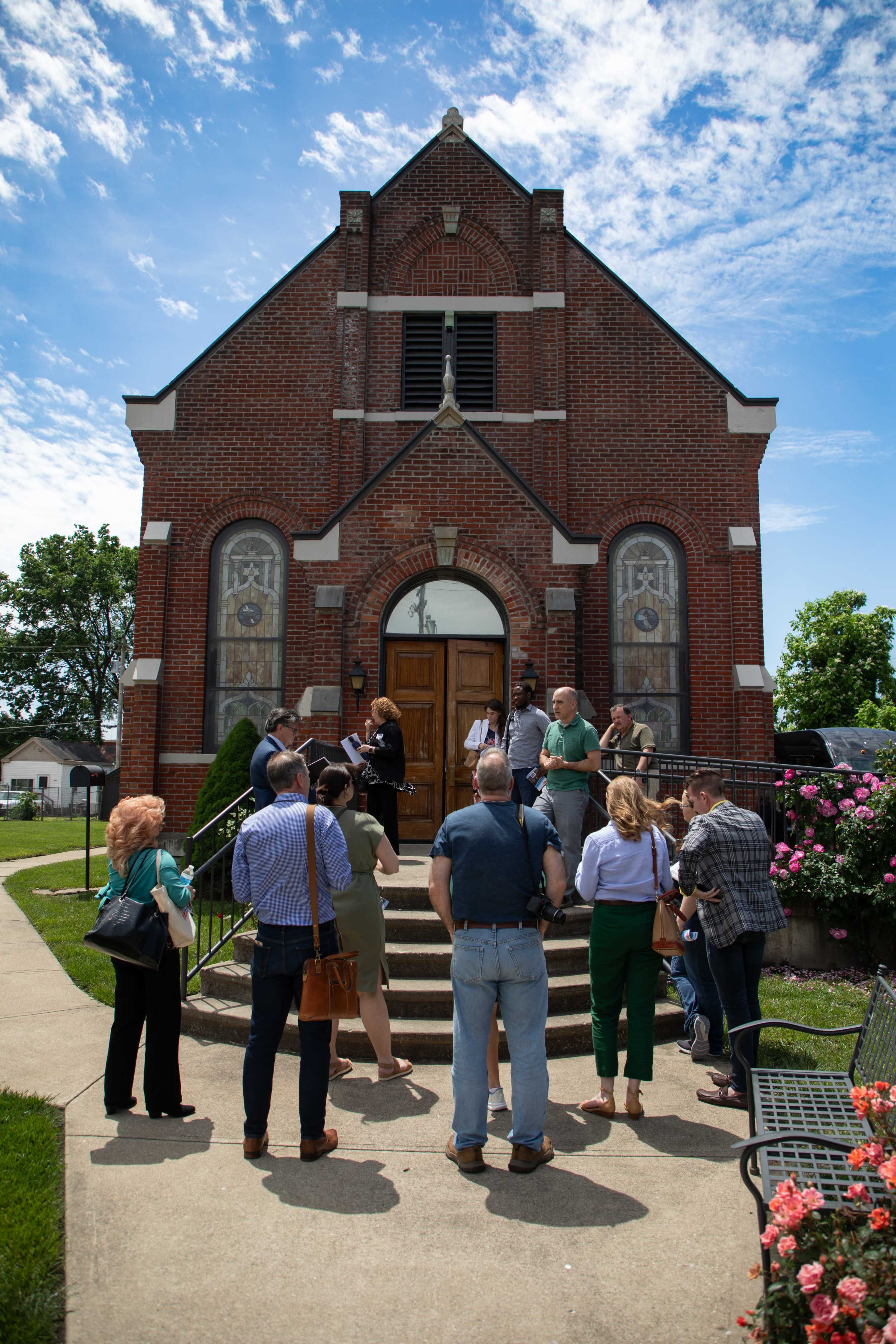 A group of people gathers in front of a brick church with decorative windows and a staircase leading to the entrance.