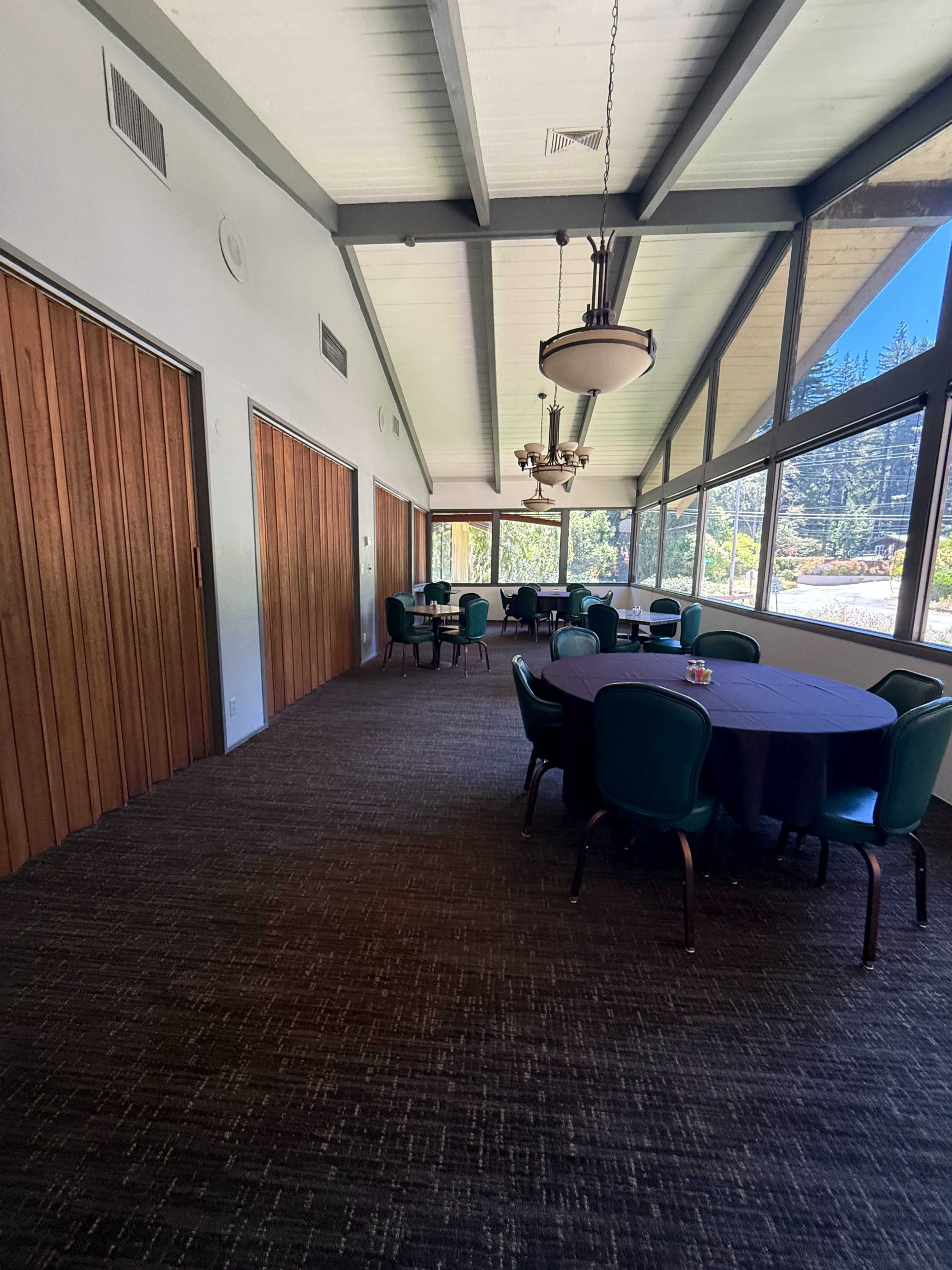 The image shows a well-lit hallway leading to a dining area with a row of tables covered in dark tablecloths and surrounded by green chairs.