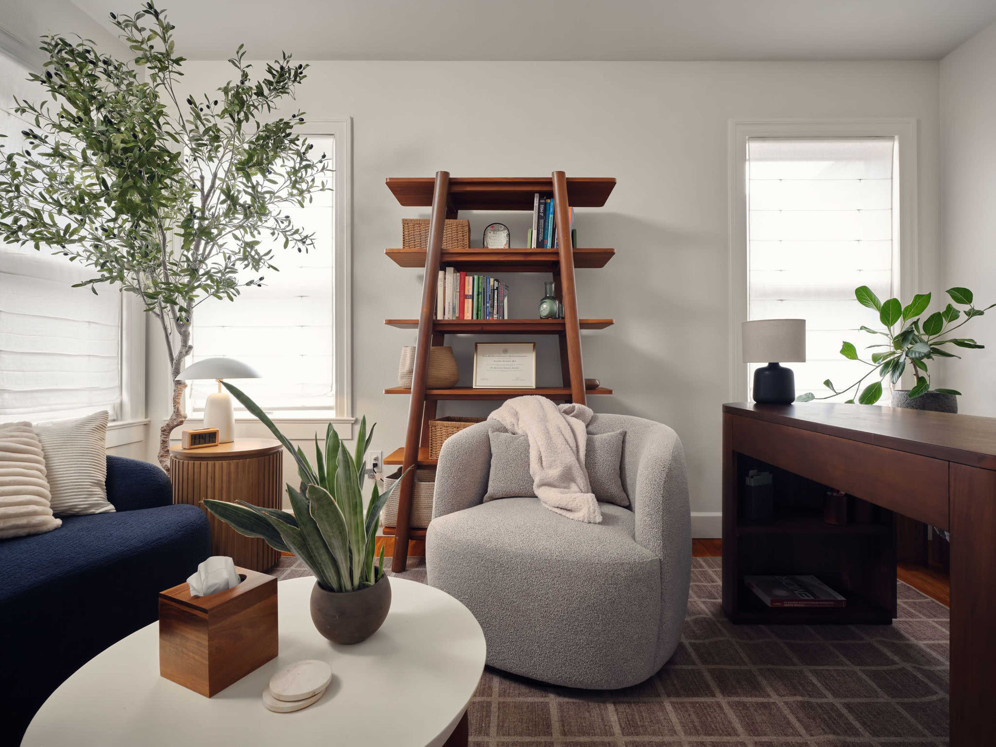 The image shows a modern living space featuring a cozy armchair, a wooden bookshelf with books and decorative items, a small coffee table with plants, and a desk against the wall.