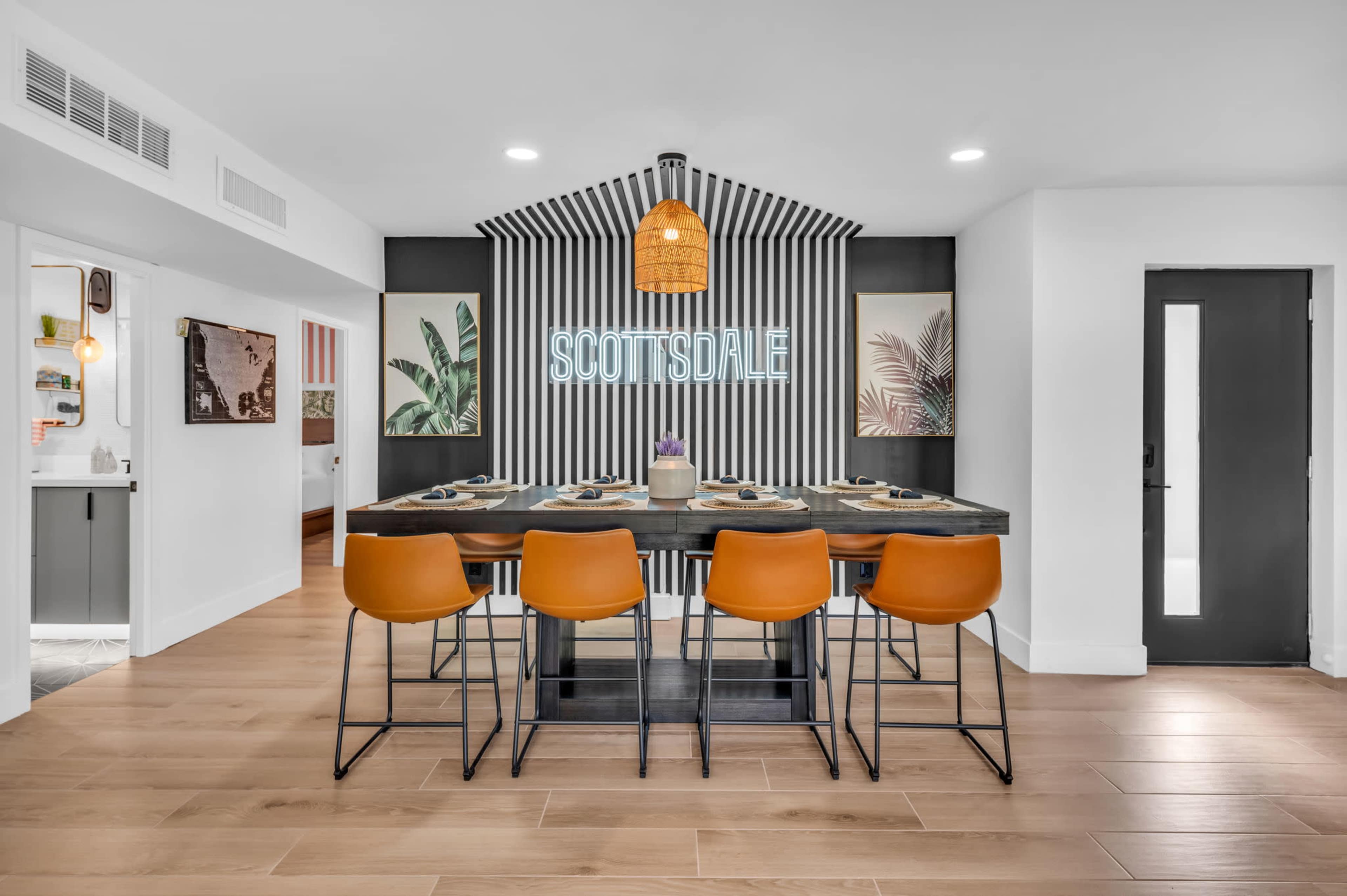 A modern dining area features a long black table with orange stools, a neon "SCOTTSDALE" sign, and palm-themed artwork against striped walls.