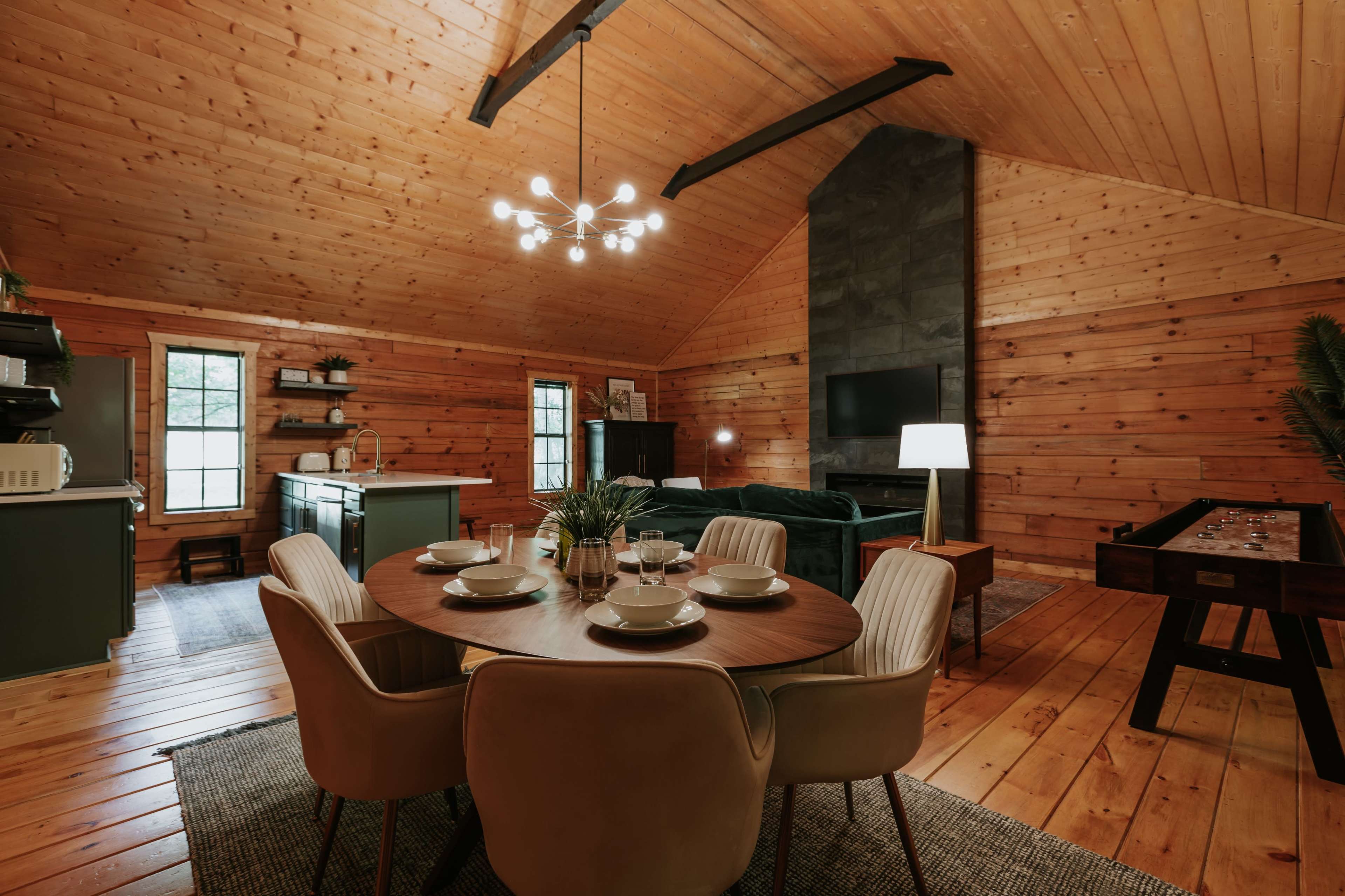 A cozy wooden cabin interior with a round dining table set for four, a living area featuring a dark sofa and a fireplace, and a modern kitchen in the background.