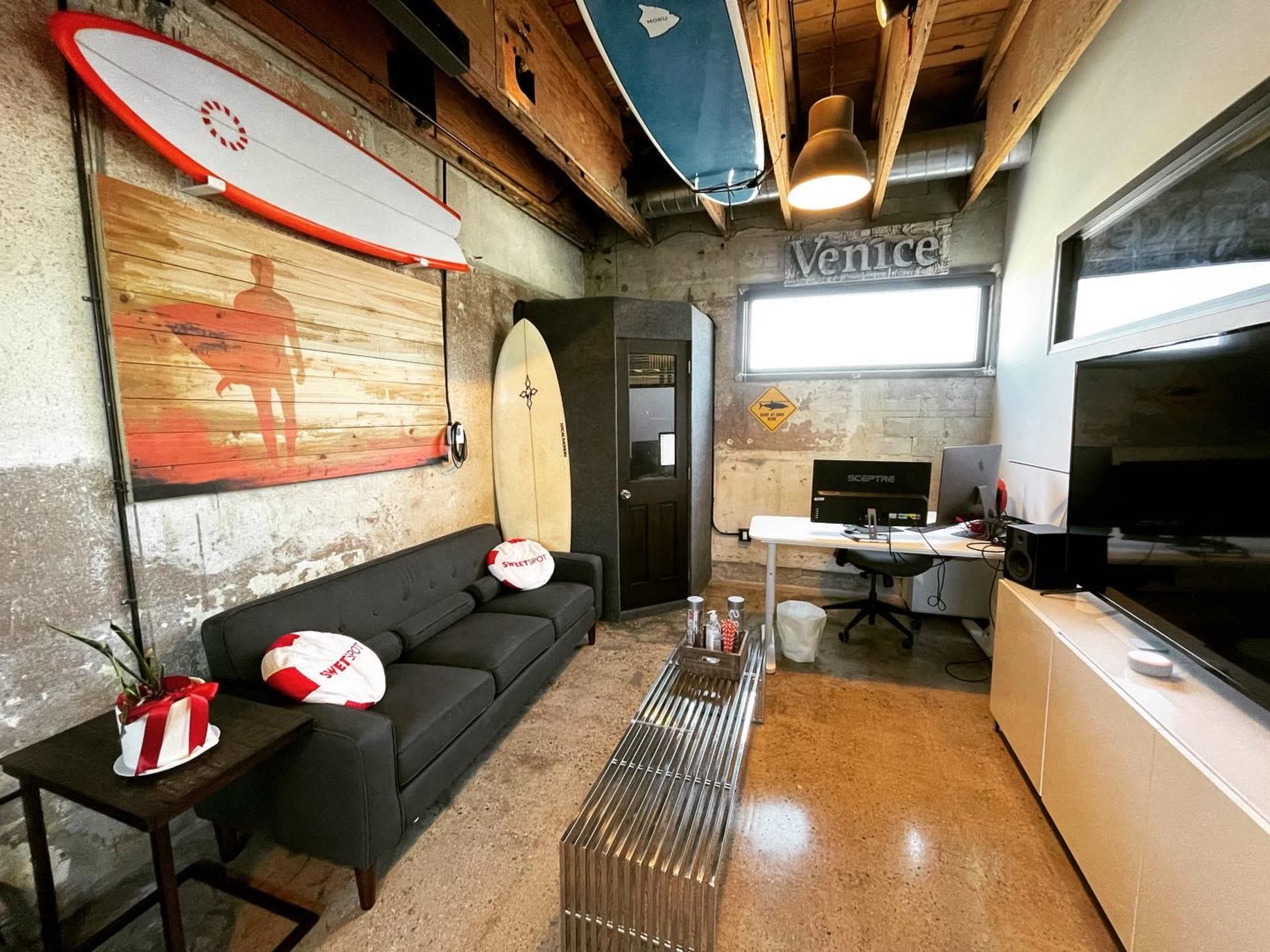 A contemporary workspace with a gray sofa, a surfboard mounted on the wall, and a desk setup with a computer and television in a minimalist room with exposed concrete walls.
