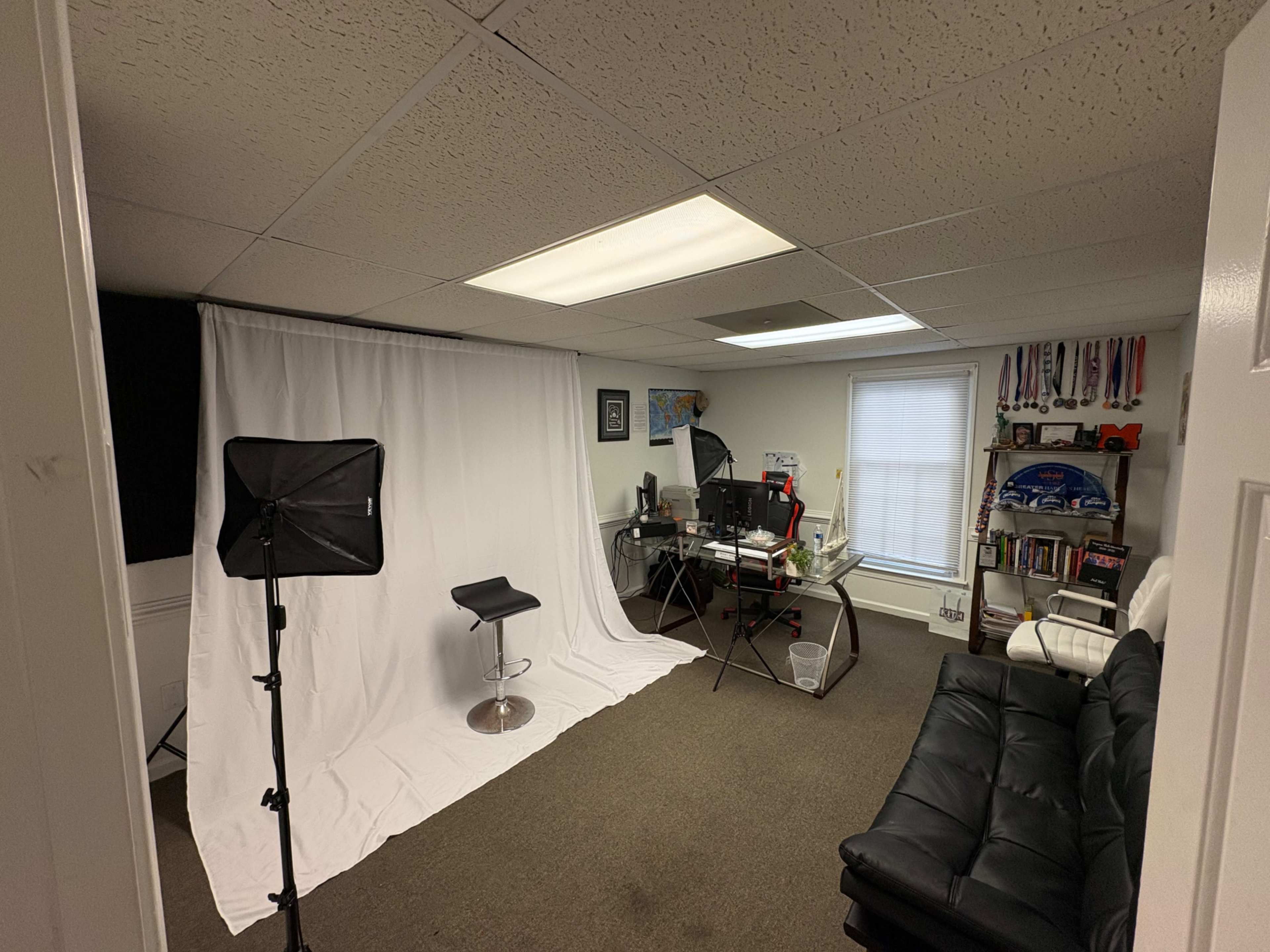 The image shows an office space with a white backdrop, a chair, and a desk equipped with a computer, adjacent to a black leather couch and shelves filled with books and awards.