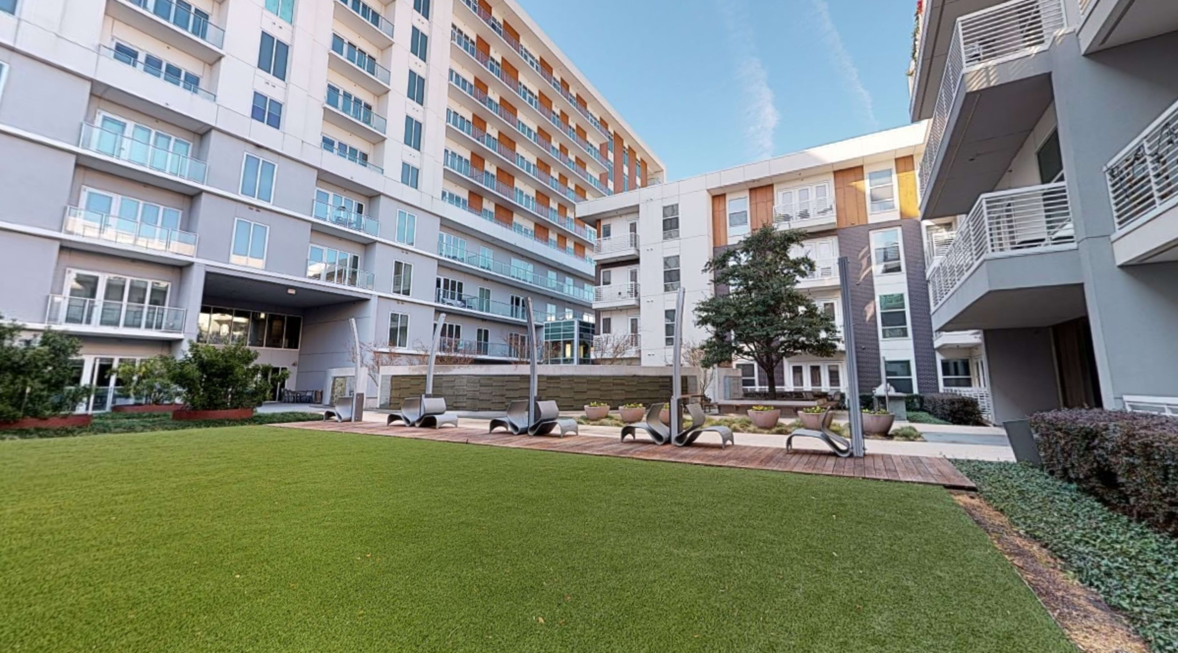 A landscaped courtyard area featuring artificial grass, seating arrangements, and several modern apartment buildings in the background.