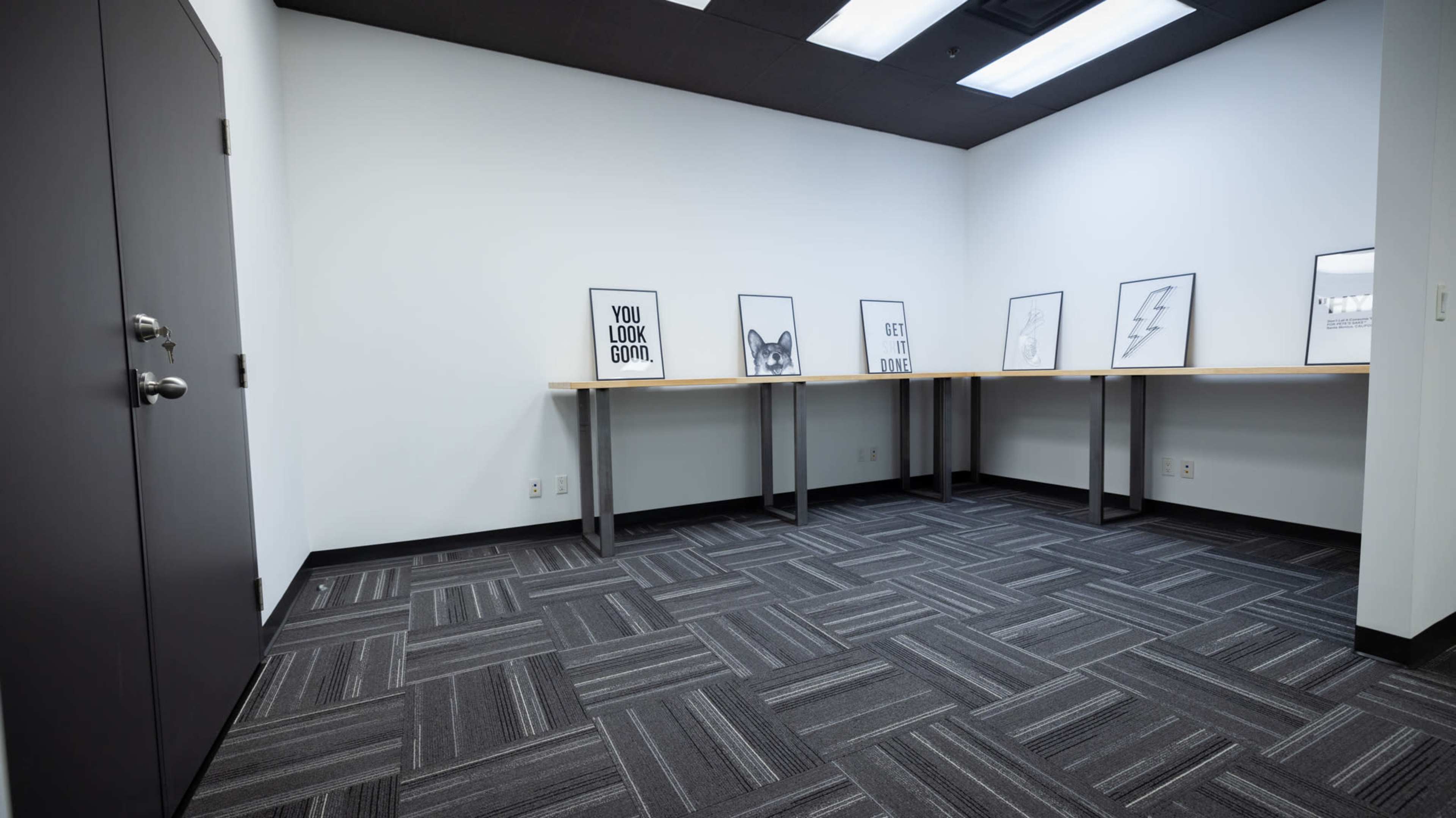 A minimalistic room with gray carpet, white walls, and several framed prints displayed on tables against one wall.