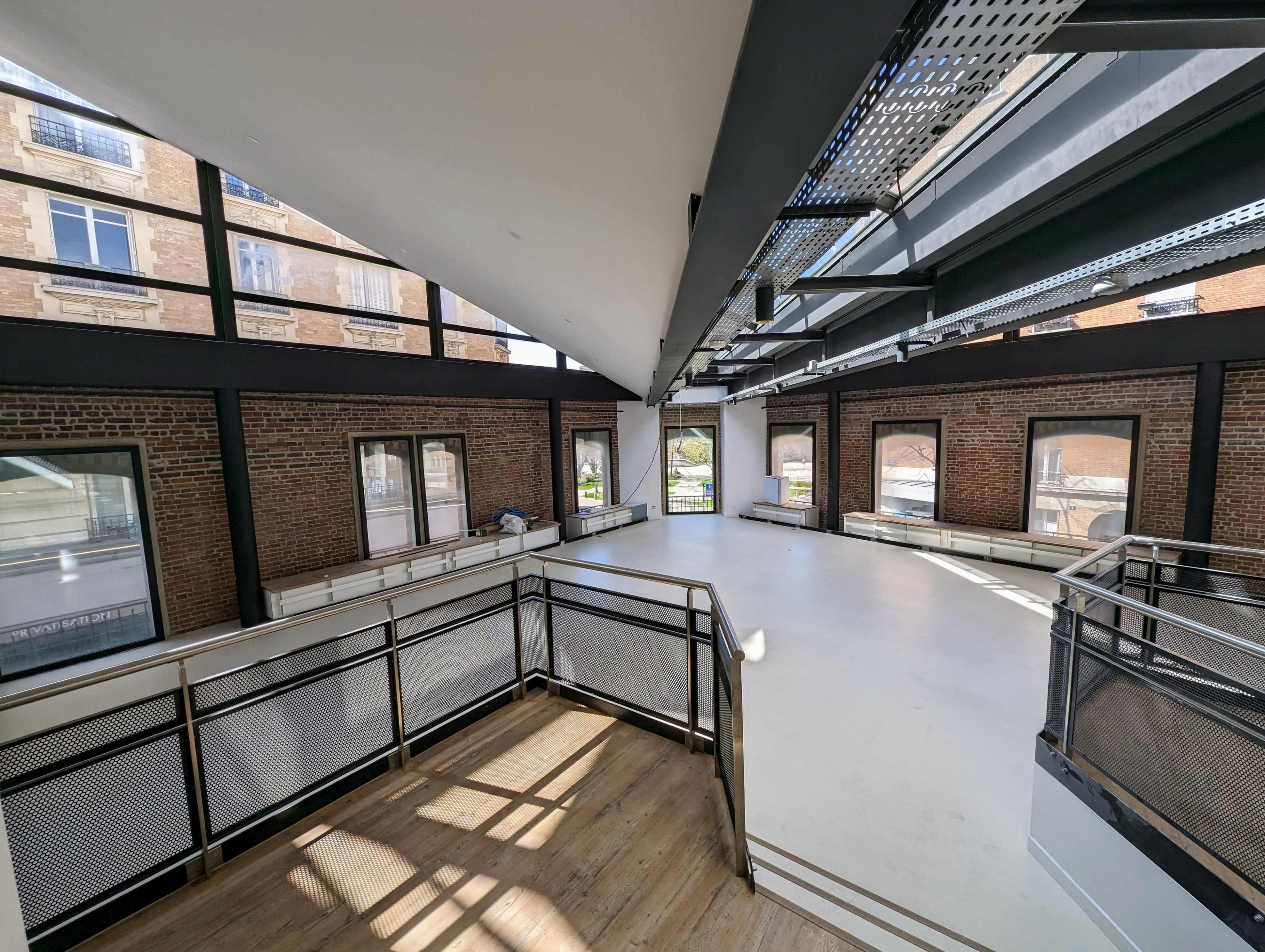 An empty room with large windows, exposed brick walls, and a mix of wooden and concrete flooring.