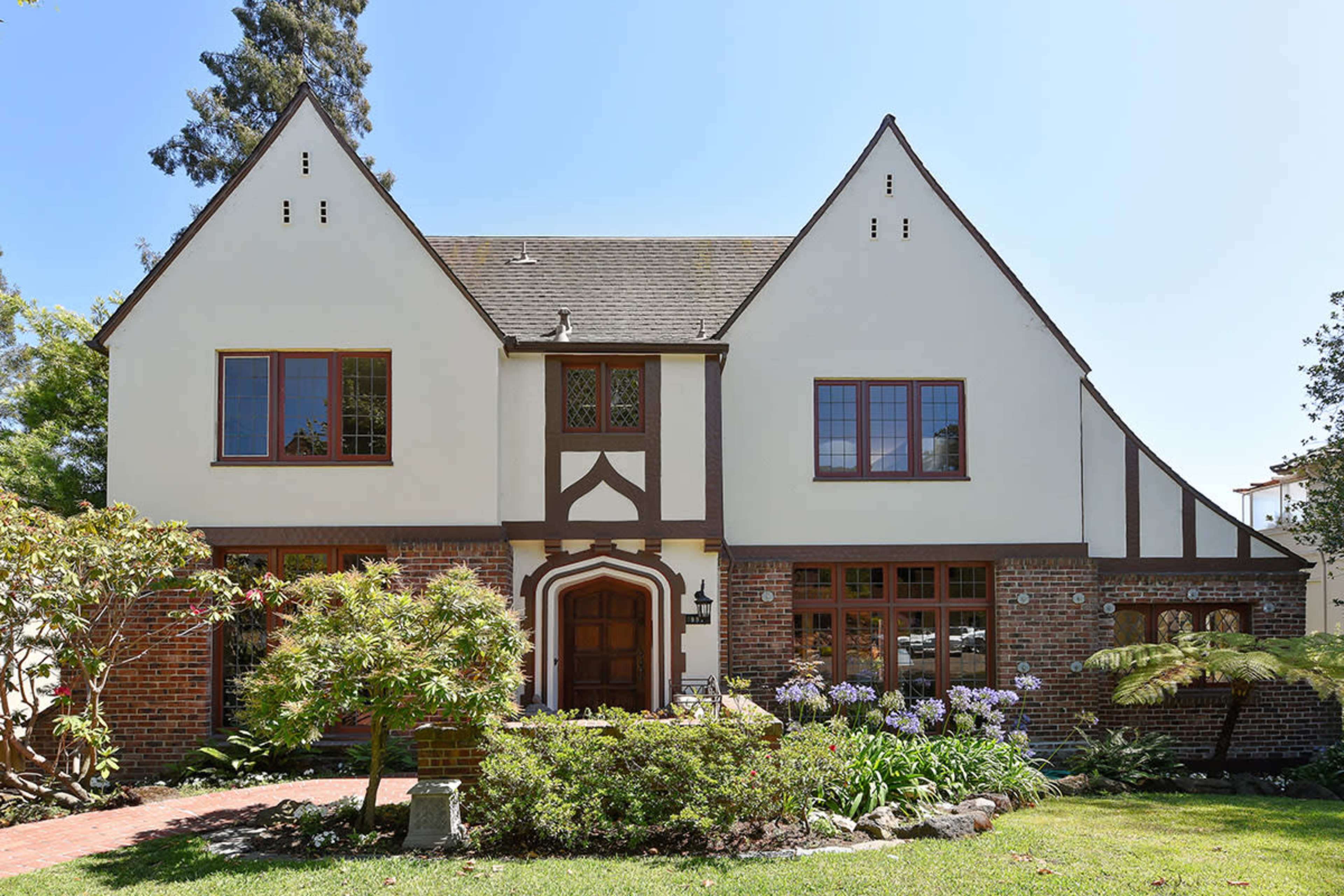The image shows a two-story Tudor-style house with a brick base, white stucco upper walls, and a landscaped front yard featuring various plants and flowers.