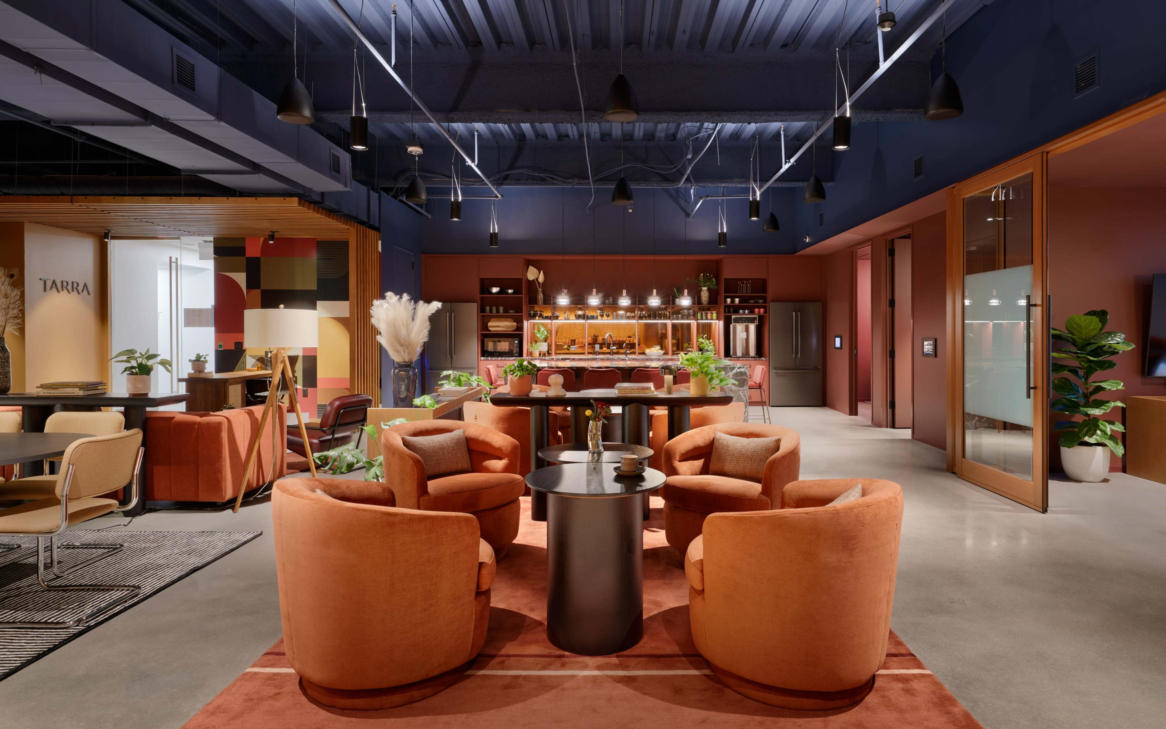 The interior features a modern lounge area with four orange armchairs surrounding a black table, set against a backdrop of wooden shelves and plants.