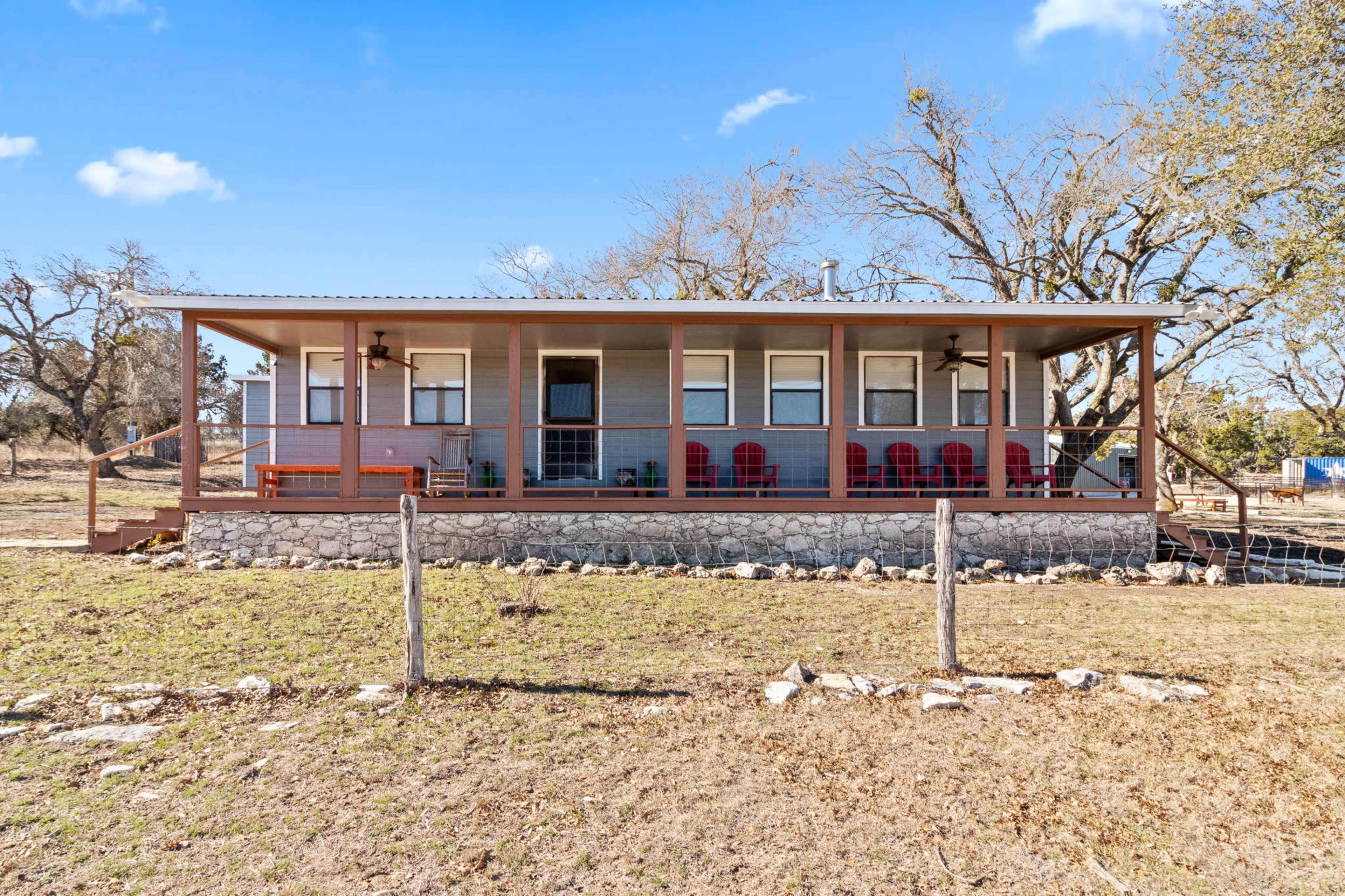 The image shows a single-story house with a covered porch, featuring several red chairs and surrounded by a grassy area and sparse trees.