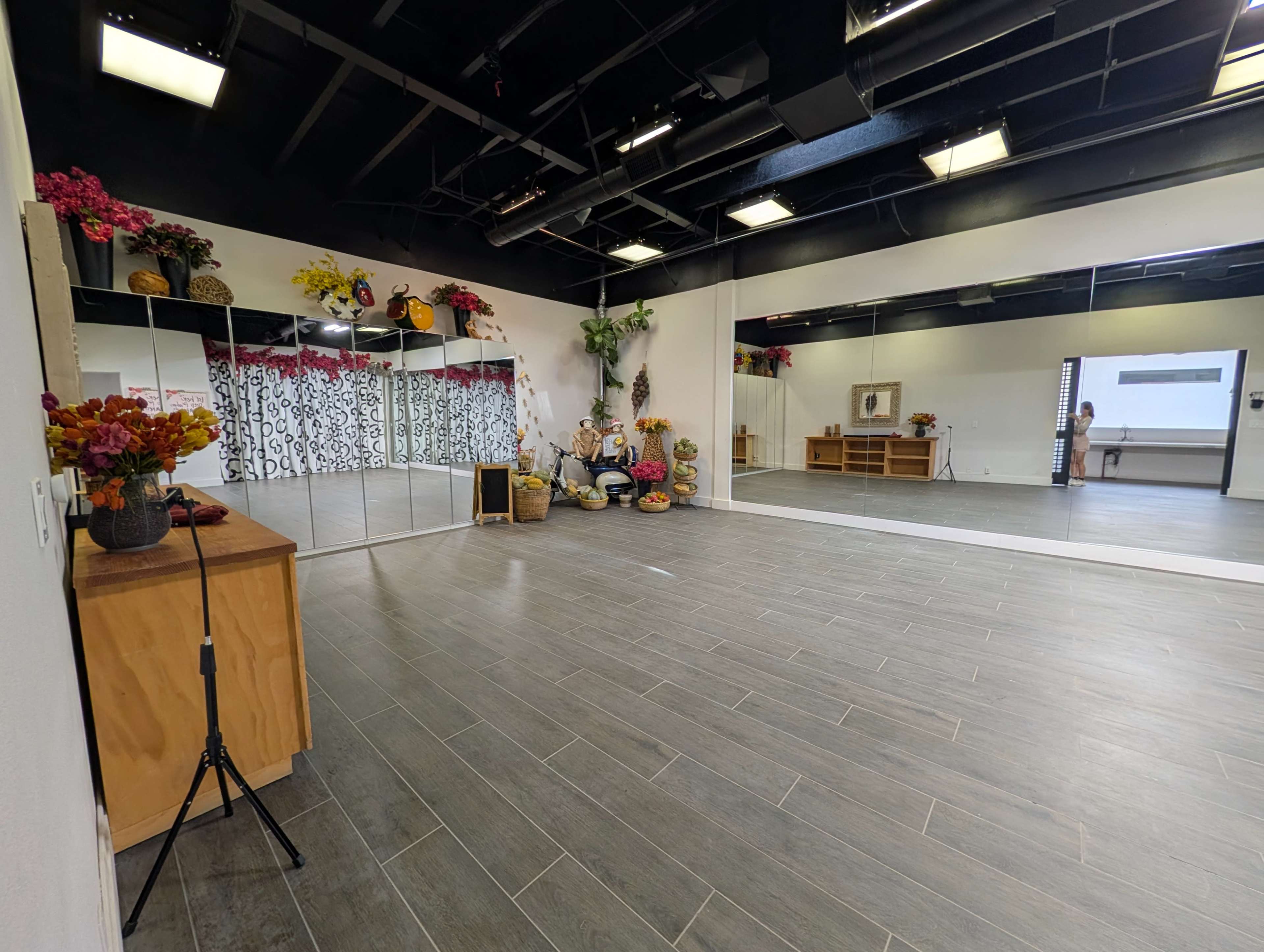 The image shows a spacious, modern studio with mirrored walls, light wooden flooring, and decorative arrangements of flowers and fruits in one corner.