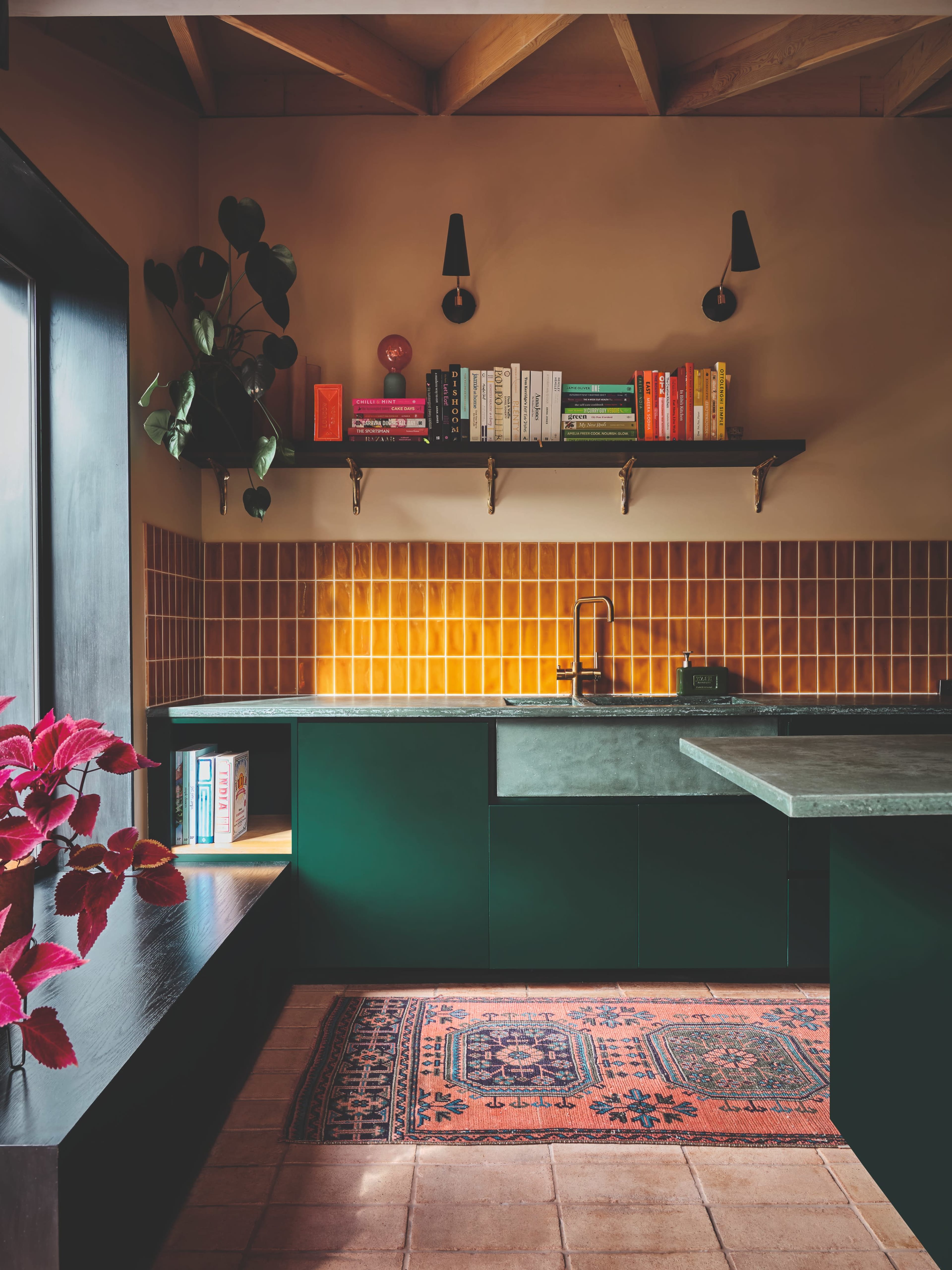 A modern kitchen features dark green cabinetry, a concrete countertop, and a wall shelf displaying colorful books, with warm lighting accentuating a tiled backsplash.