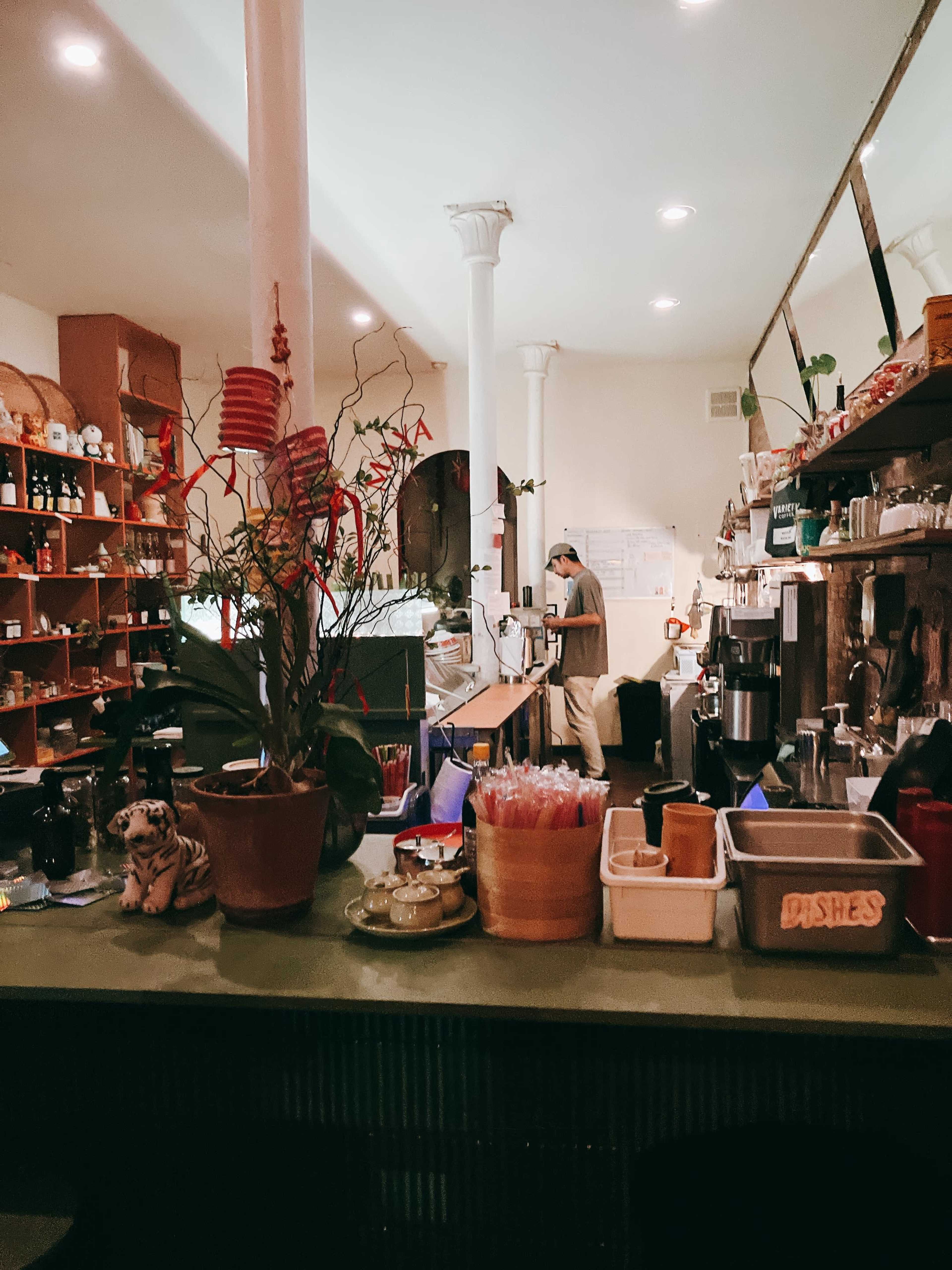 A coffee shop interior features a counter with plants and decorative items, while a barista prepares drinks in the background.