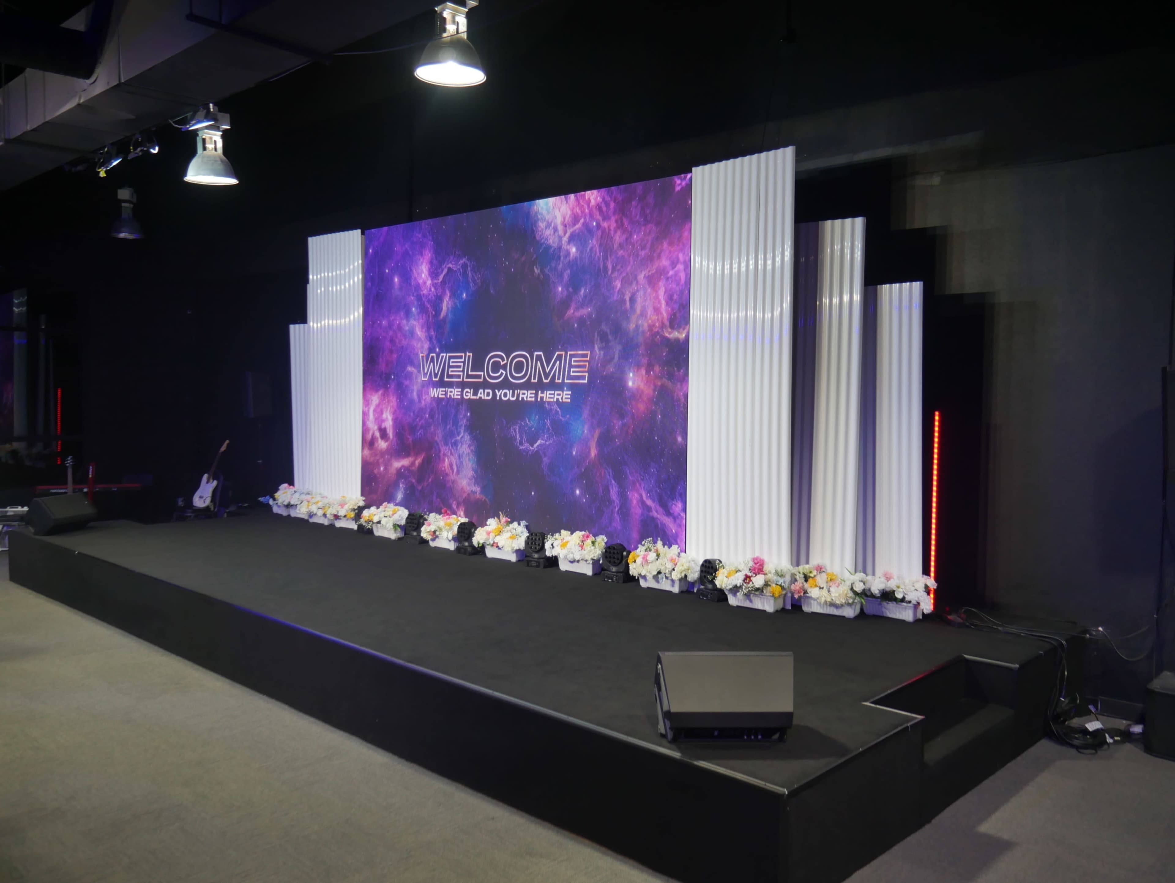 A stage is set with a colorful backdrop displaying the word "WELCOME" surrounded by floral arrangements and lighting.