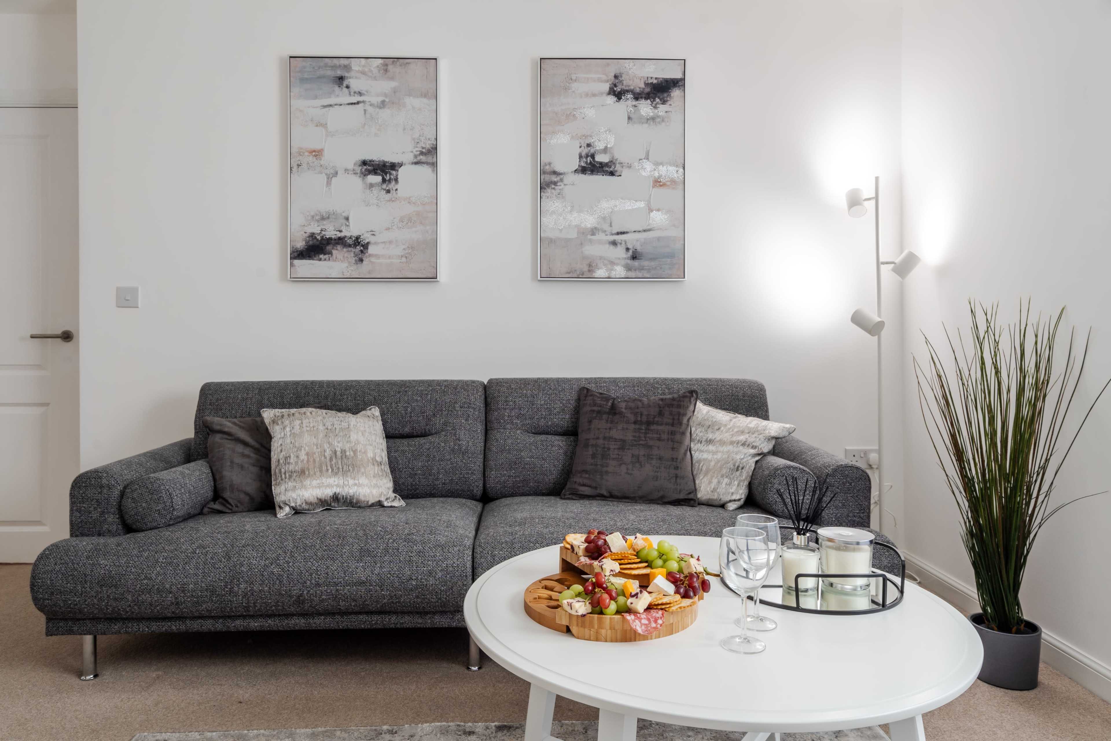 A modern living room features a gray sectional sofa and a round coffee table with a fruit platter, surrounded by artwork and a floor lamp.