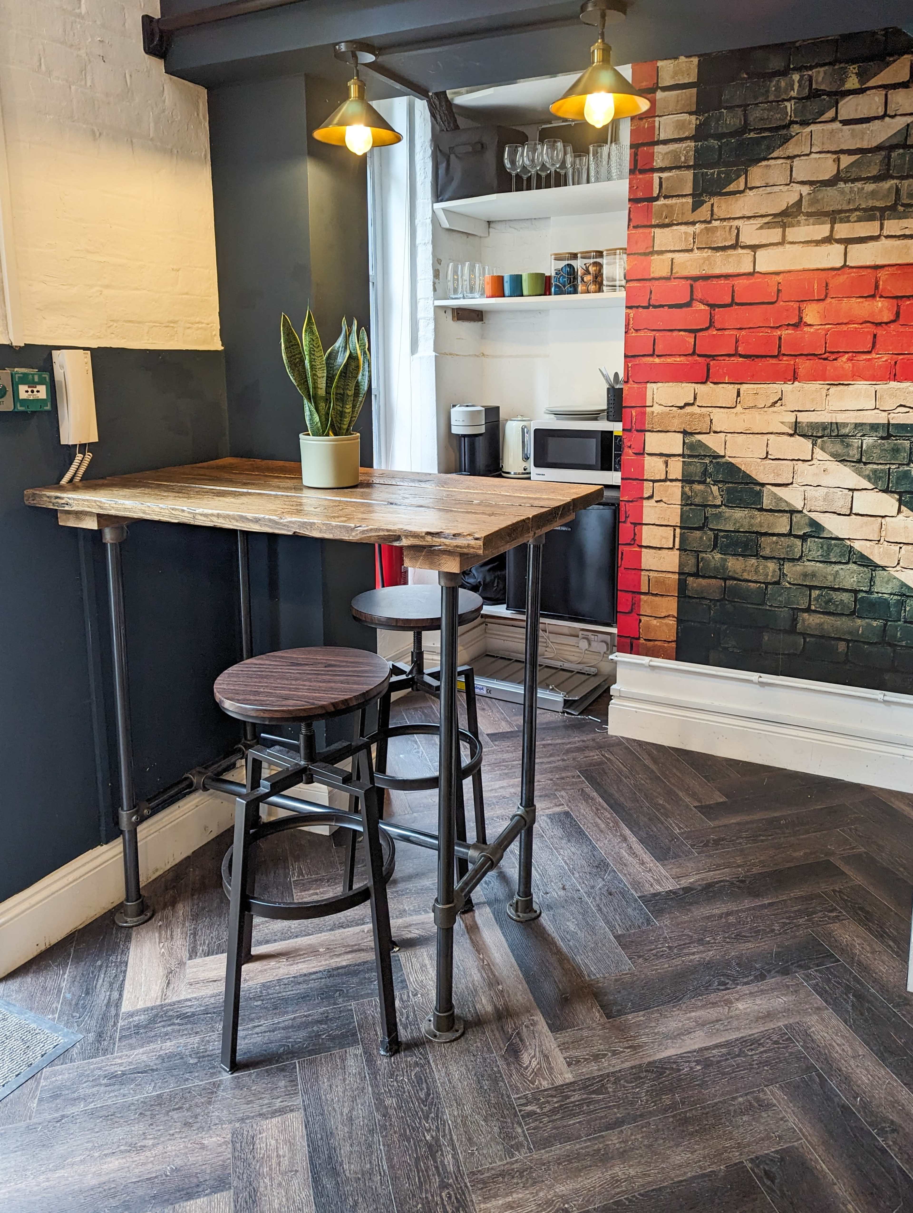 A wooden table with metal legs and two stools sits in a corner of a room featuring a Union Jack mural on one wall and modern kitchen appliances in the background.