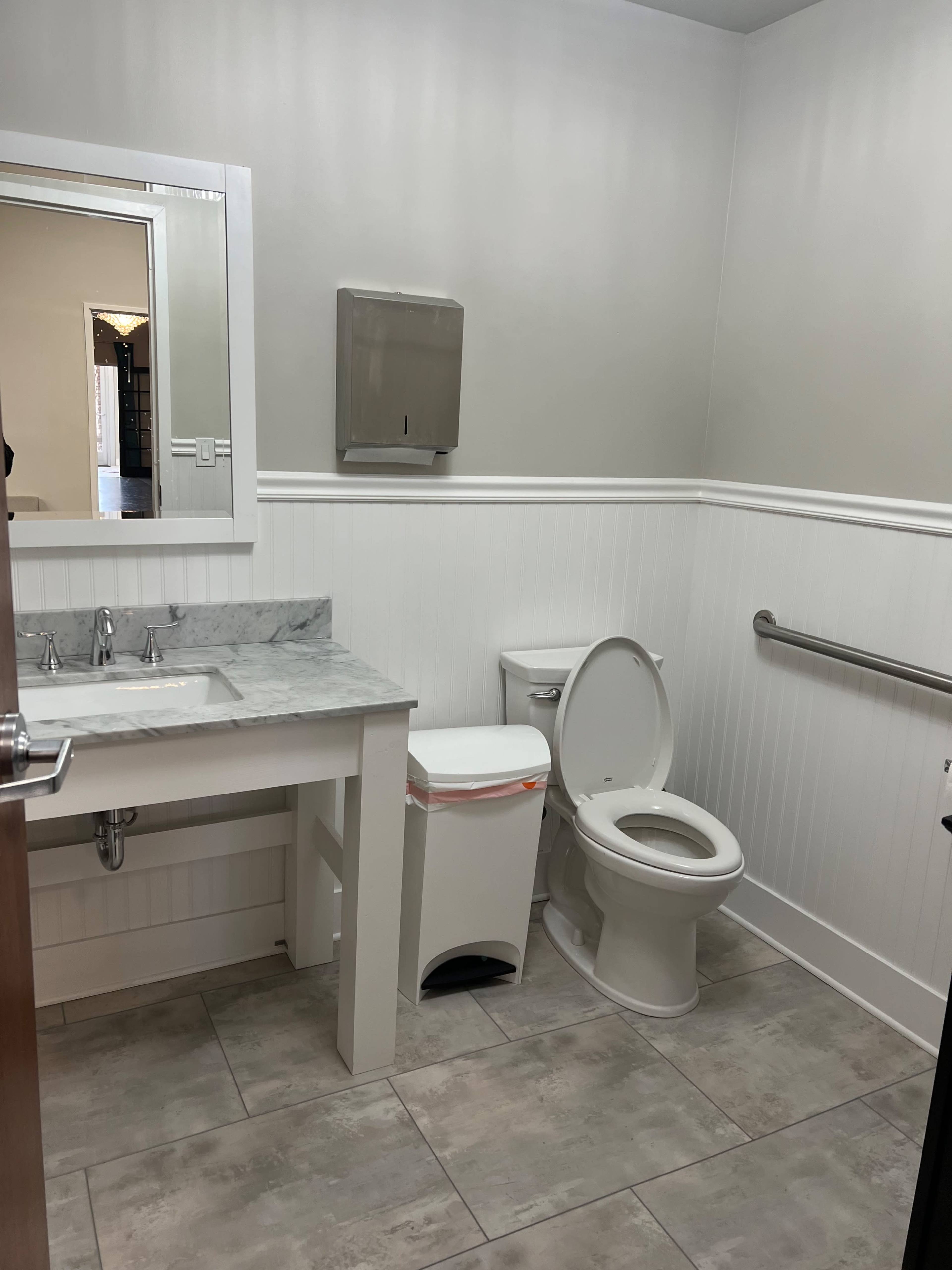 A clean bathroom with a toilet, a small sink with a marble countertop, a trash can, and a mirrored wall cabinet.