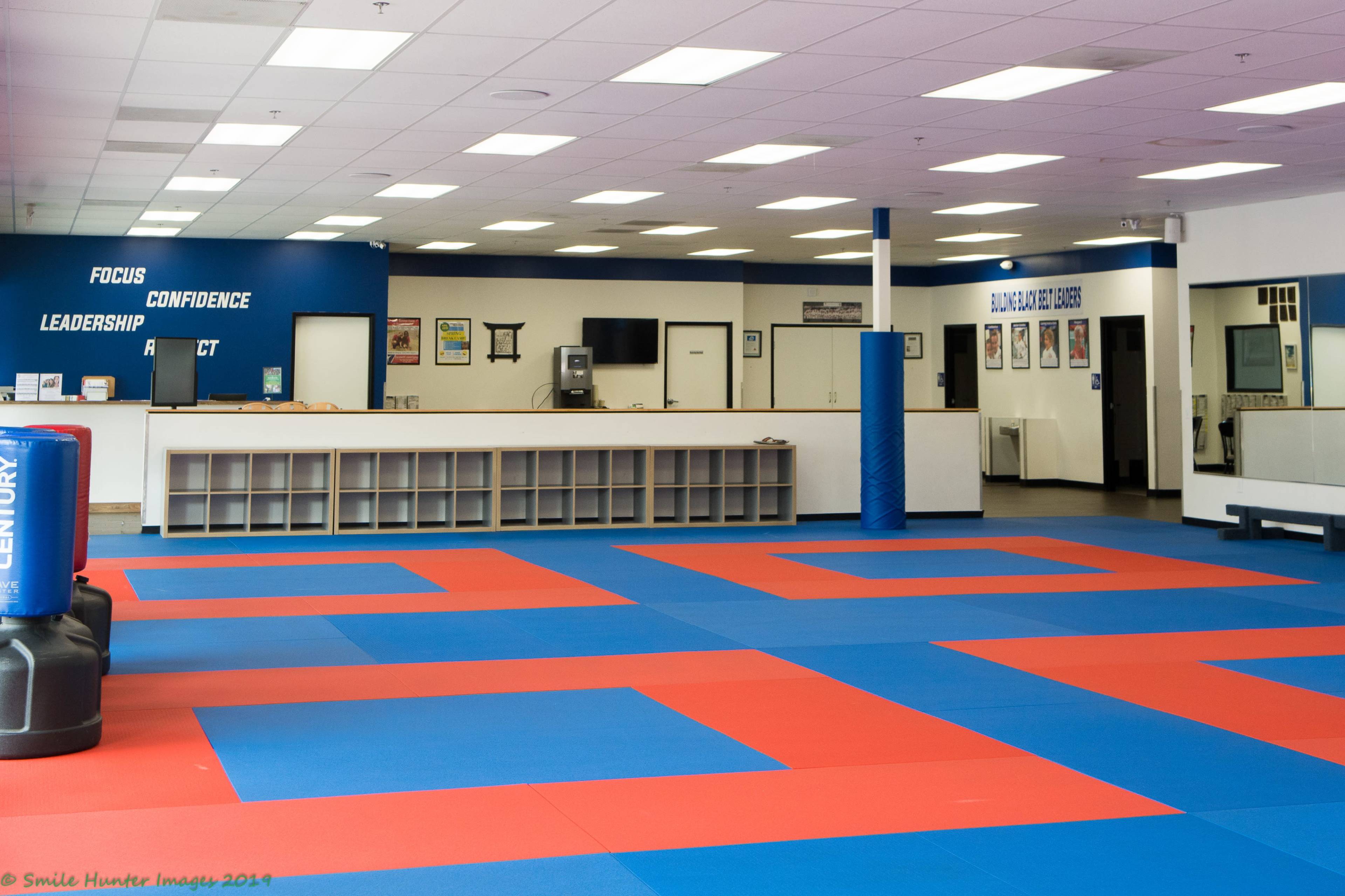 A martial arts training studio with blue and red tatami flooring, featuring an empty reception area and motivational phrases on the wall.