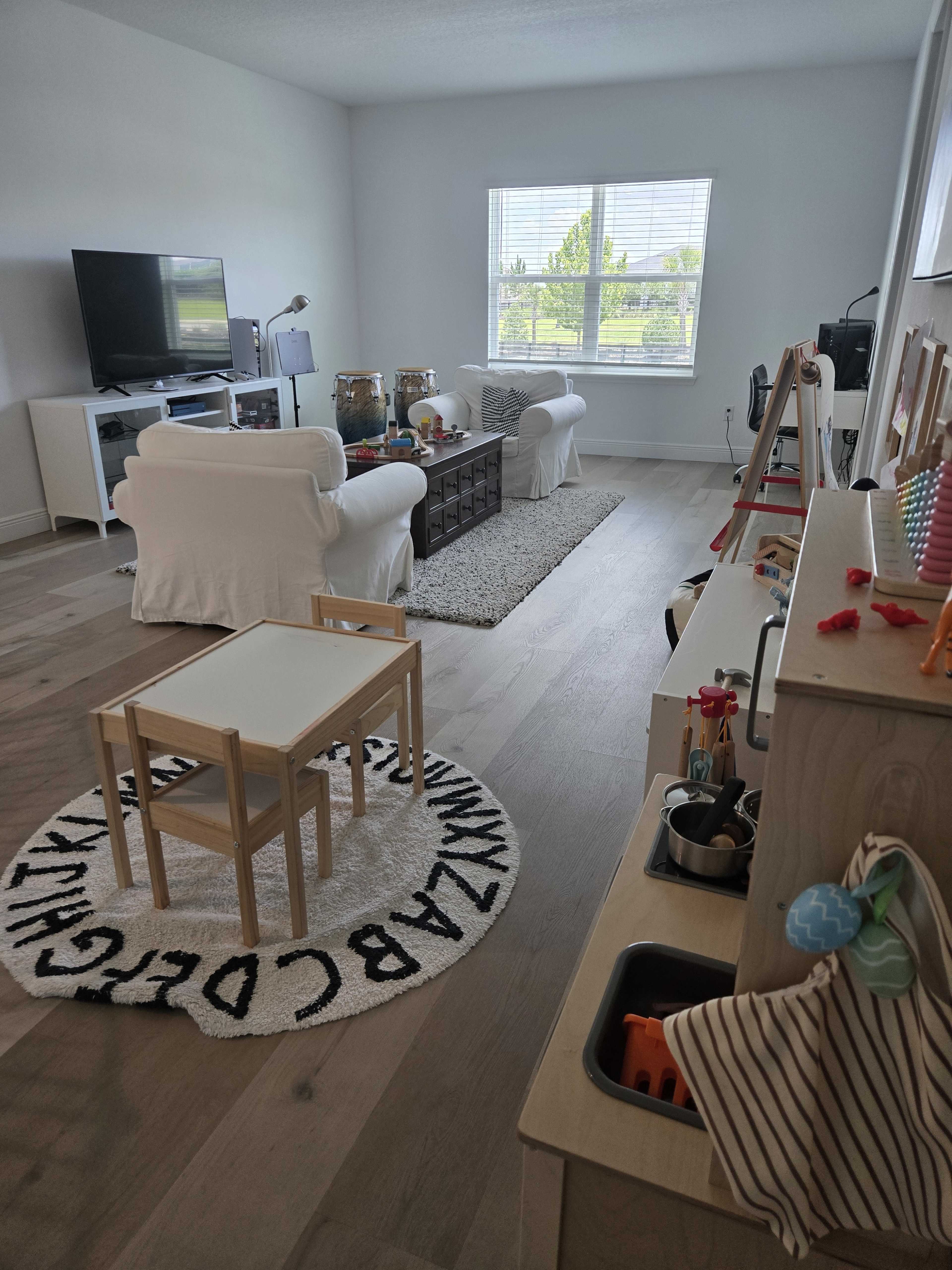 The image shows a bright living room featuring a white couch, a television, a coffee table, and children's toys arranged in a play area.