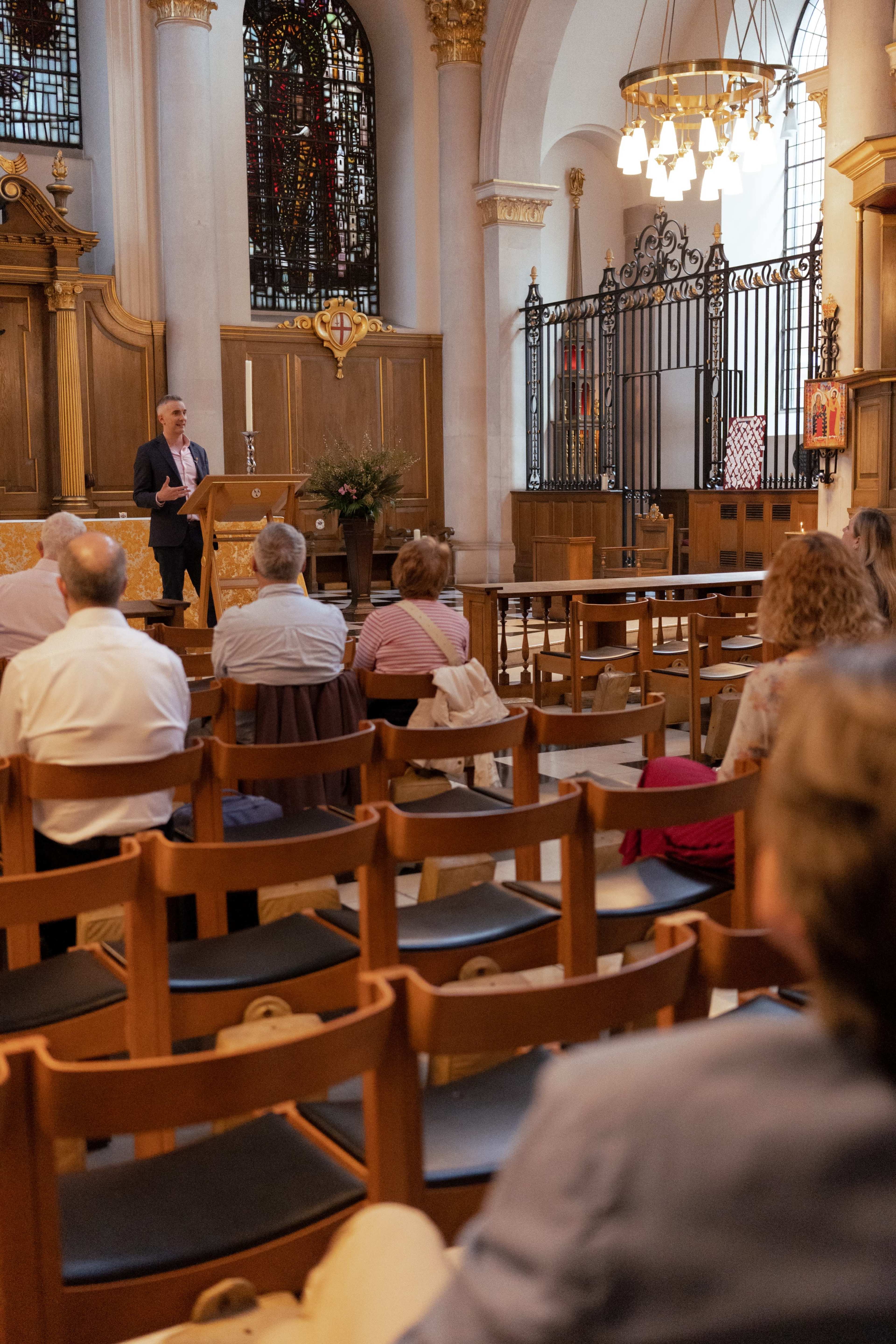A speaker stands at the front of a church hall, addressing a small audience seated in wooden chairs.