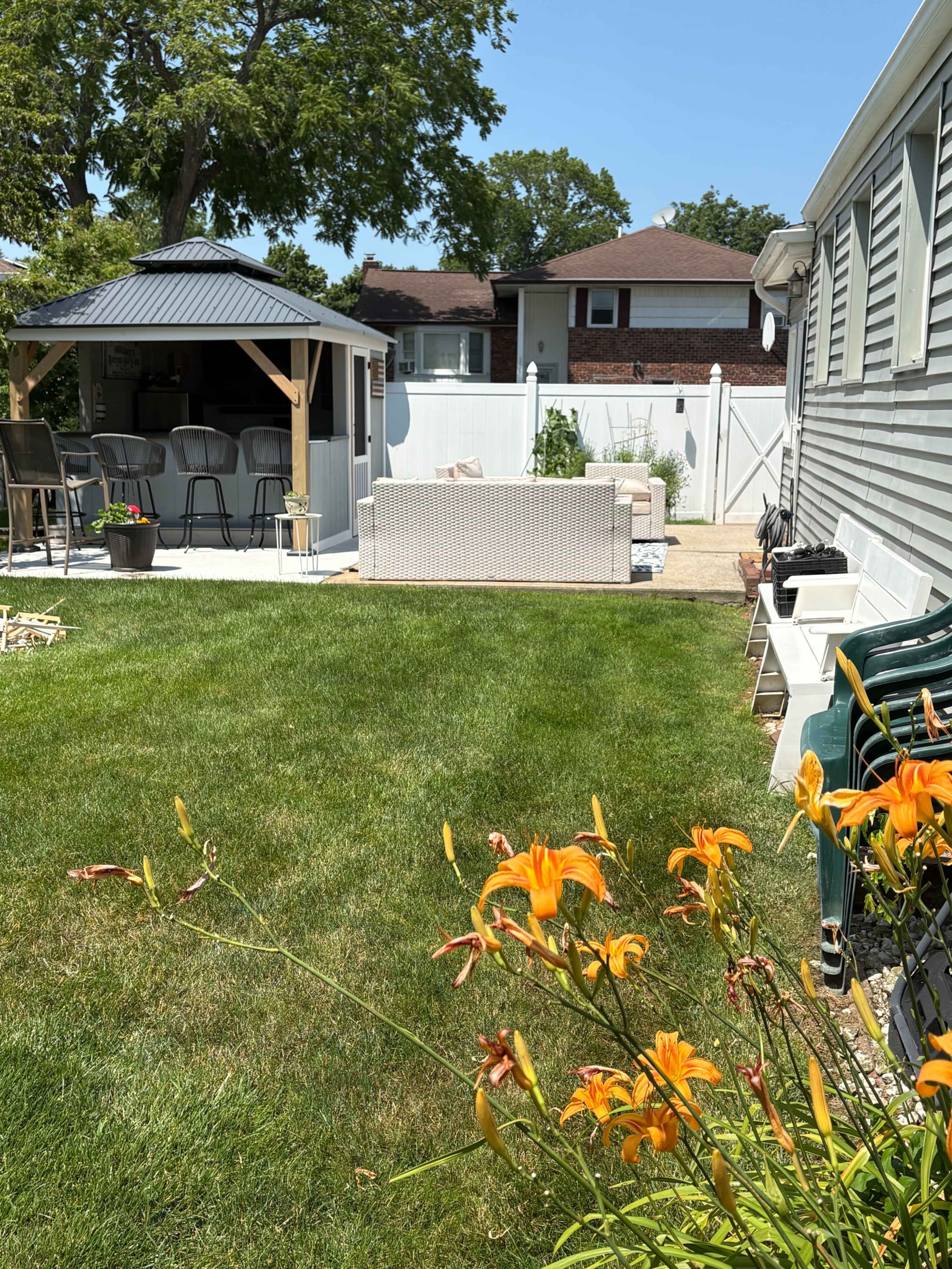 A well-maintained backyard features a gazebo with seating, a patio area with a light-colored couch, and vibrant orange flowers in the foreground.