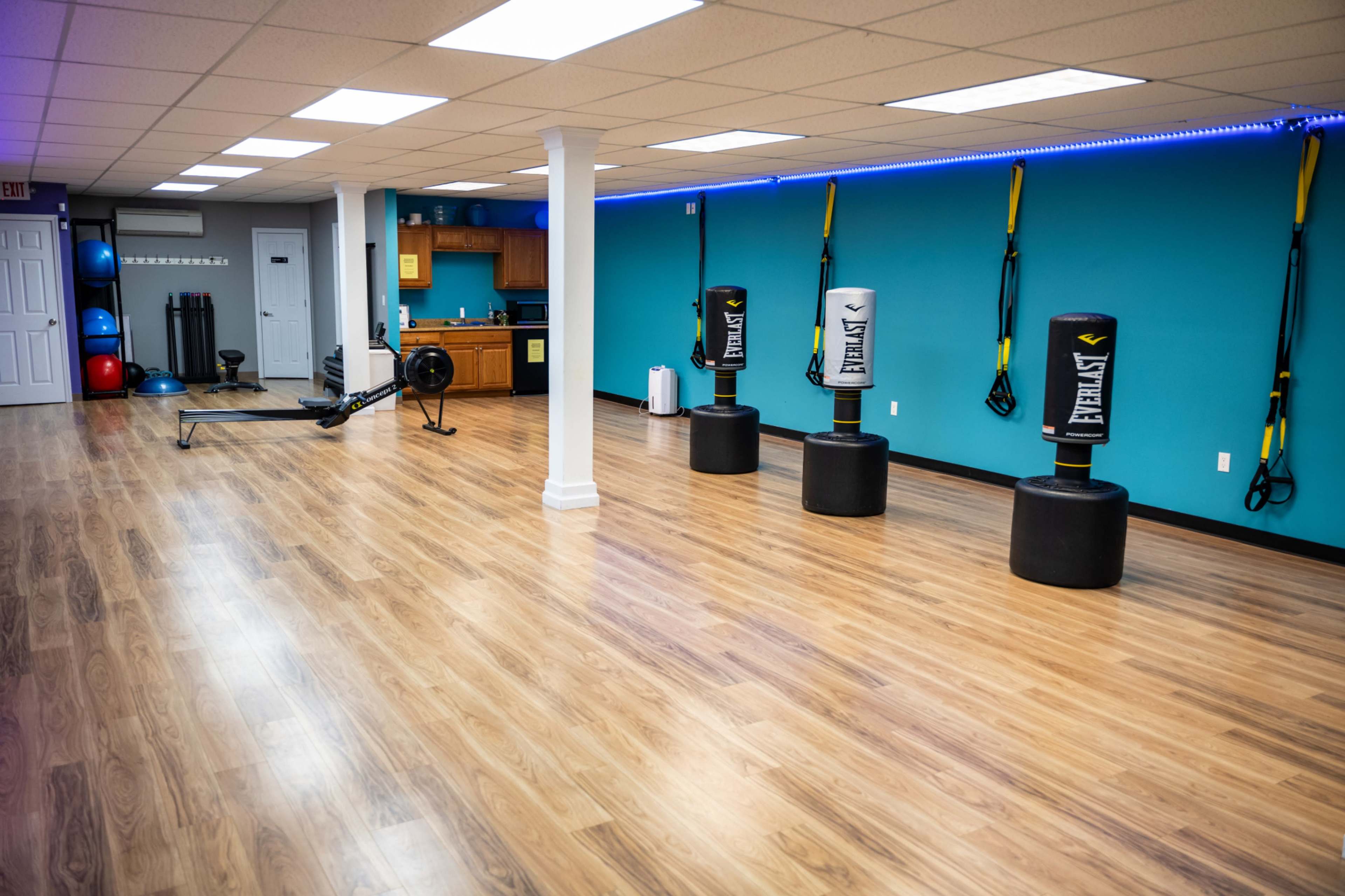 A spacious fitness studio with wooden floors, blue walls, and various equipment including punching bags, exercise balls, and a kitchen area.