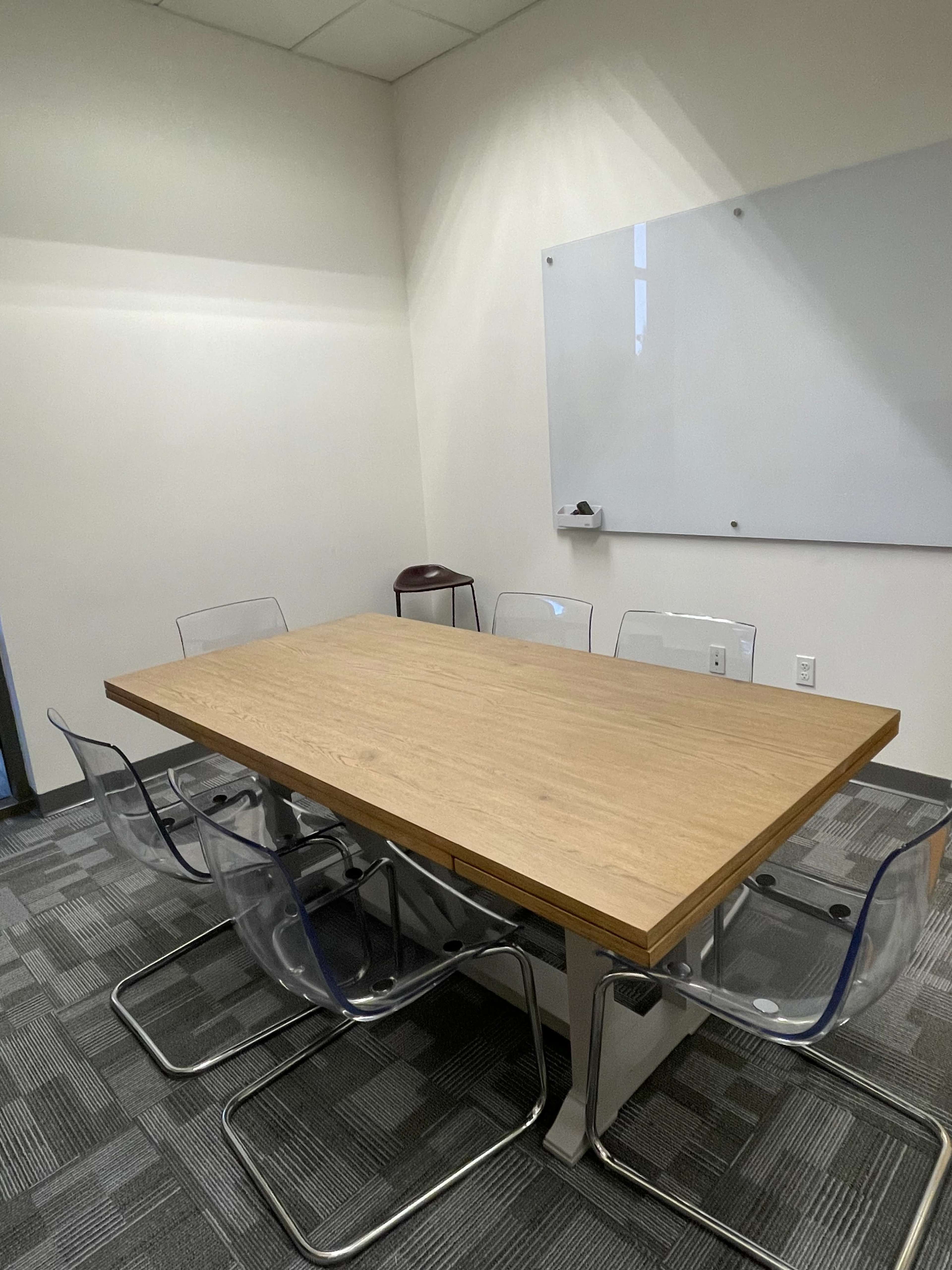 The image shows a small conference room with a large wooden table surrounded by six clear plastic chairs and a whiteboard on the wall.