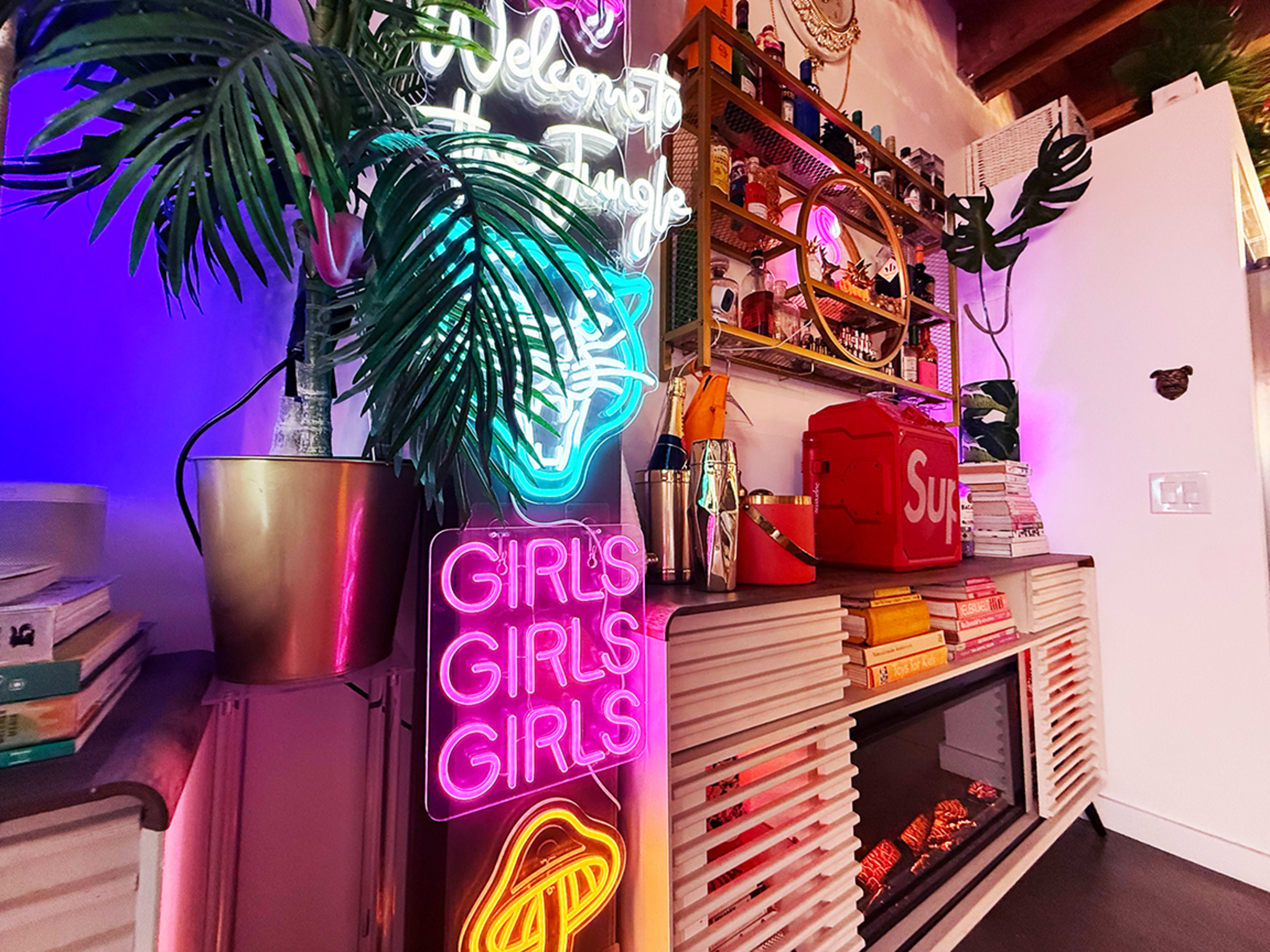 The image shows a vibrant interior scene featuring neon signs, colorful decor, and a bookshelf decorated with various items, including a red bag and houseplants.