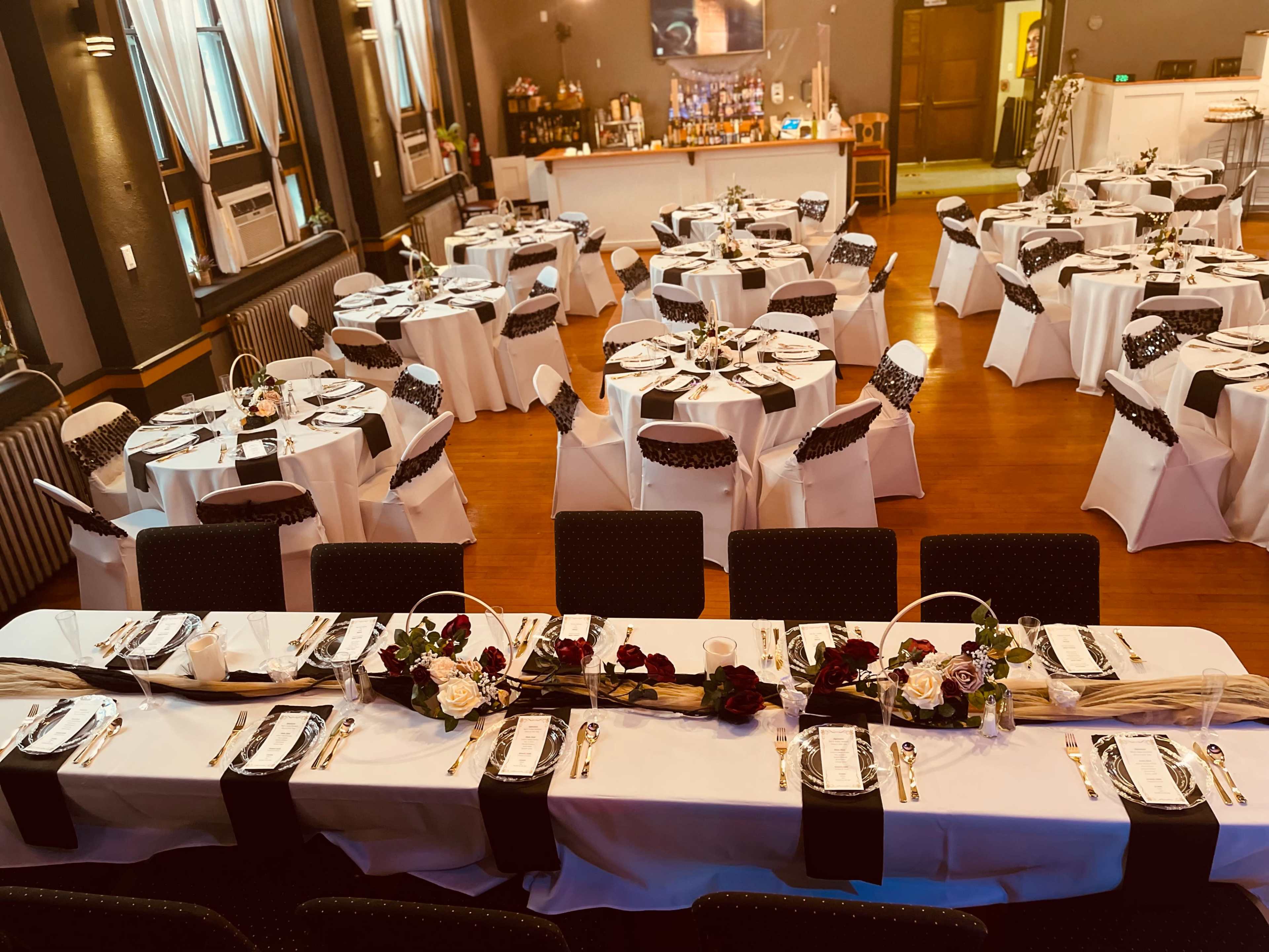 The image shows a banquet hall set up for an event, featuring neatly arranged tables covered with white tablecloths and dark chair sashes, adorned with floral centerpieces.