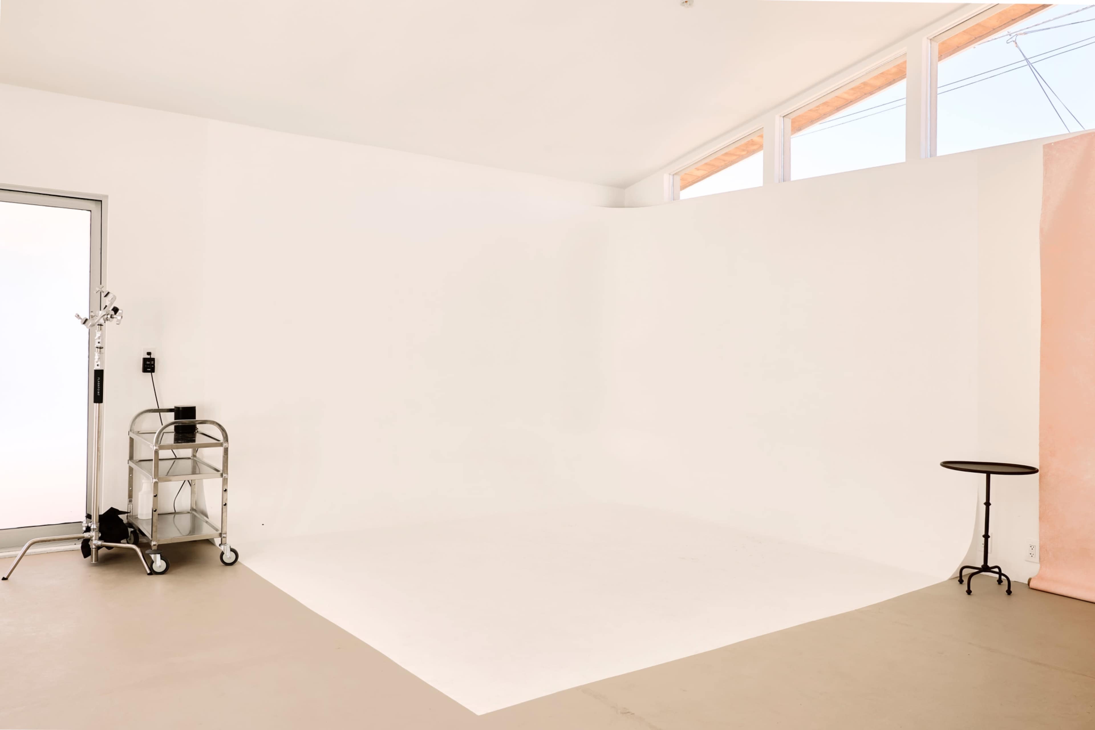 The image shows a minimalistic, well-lit studio space with white walls, a small black table, and a cart with equipment.