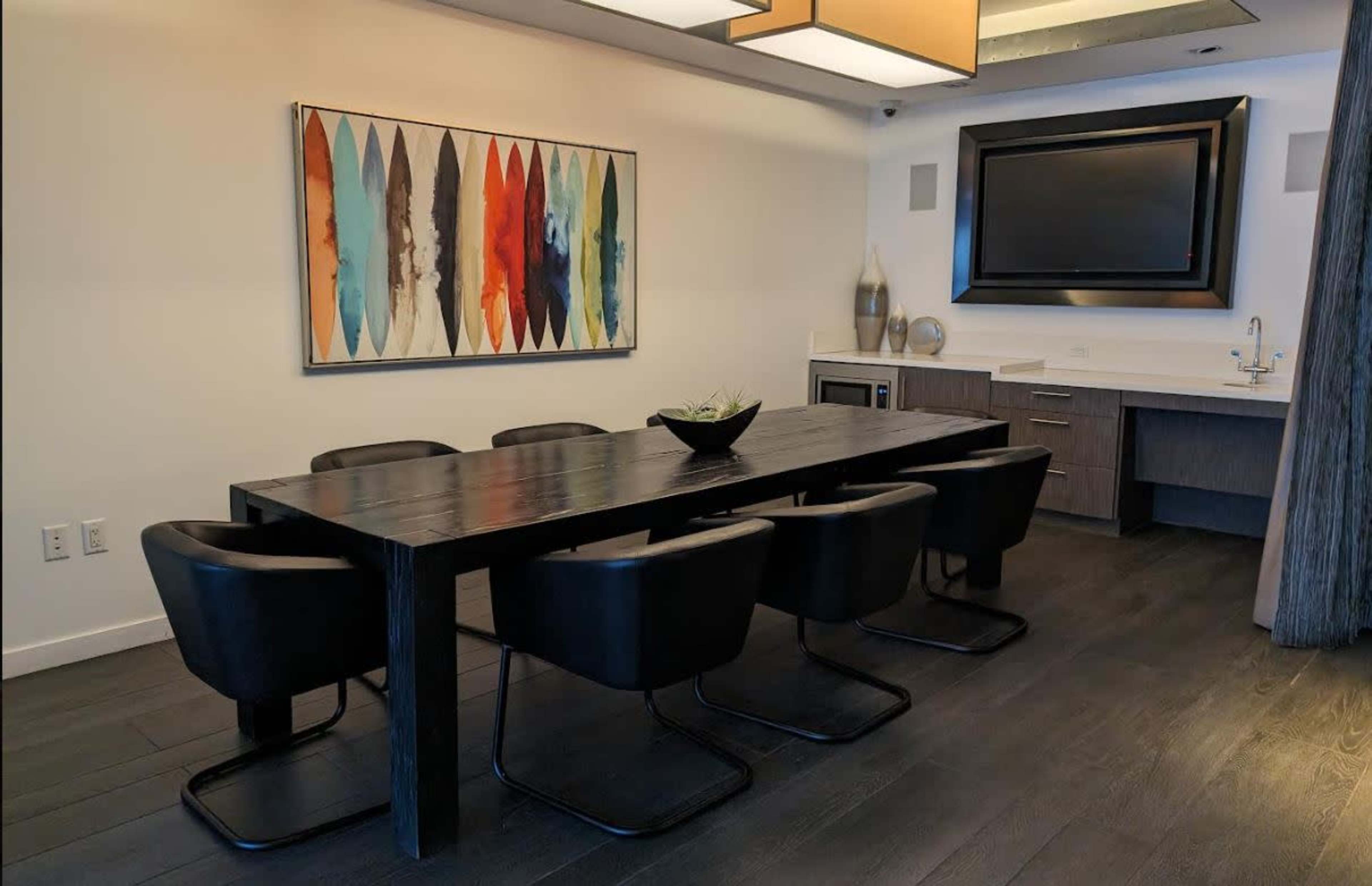 A dining area features a long black table surrounded by six black chairs, with a colorful abstract painting on the wall and a kitchenette in the background.