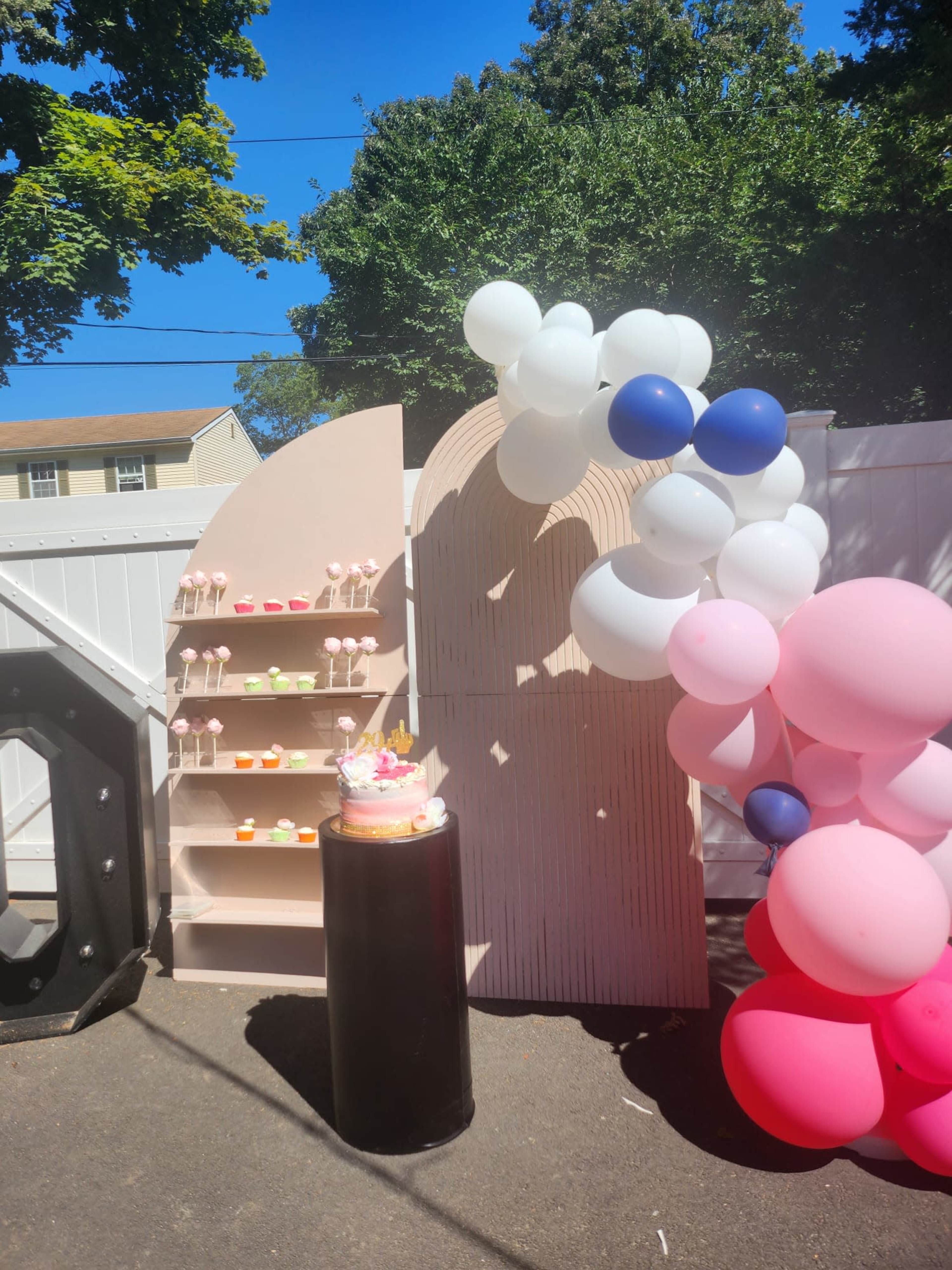 The scene features a decorative outdoor setup with a tiered display of colorful treats, a pink cake on a pedestal, and a backdrop of balloons in various colors.