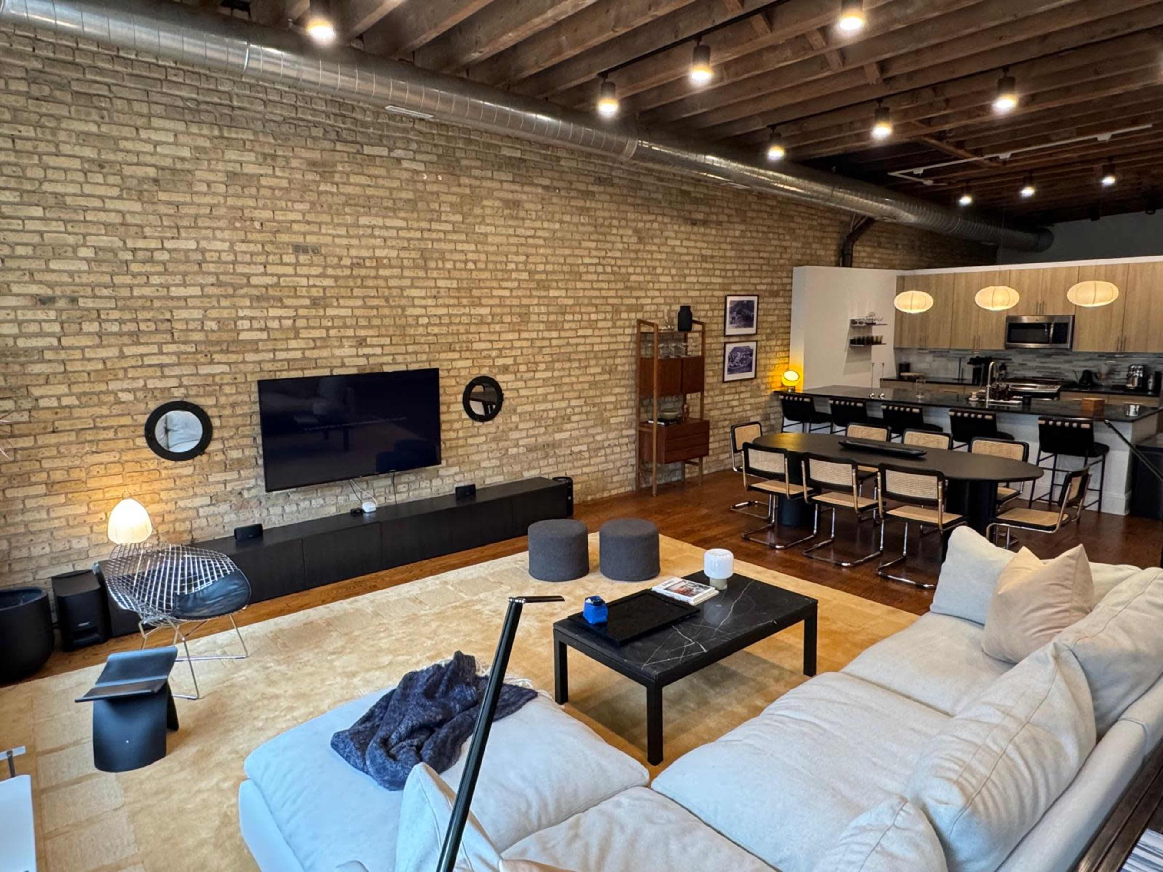The image shows a modern living room with exposed brick walls, a large sectional sofa, a coffee table, and an open kitchen area with dining seating in the background.