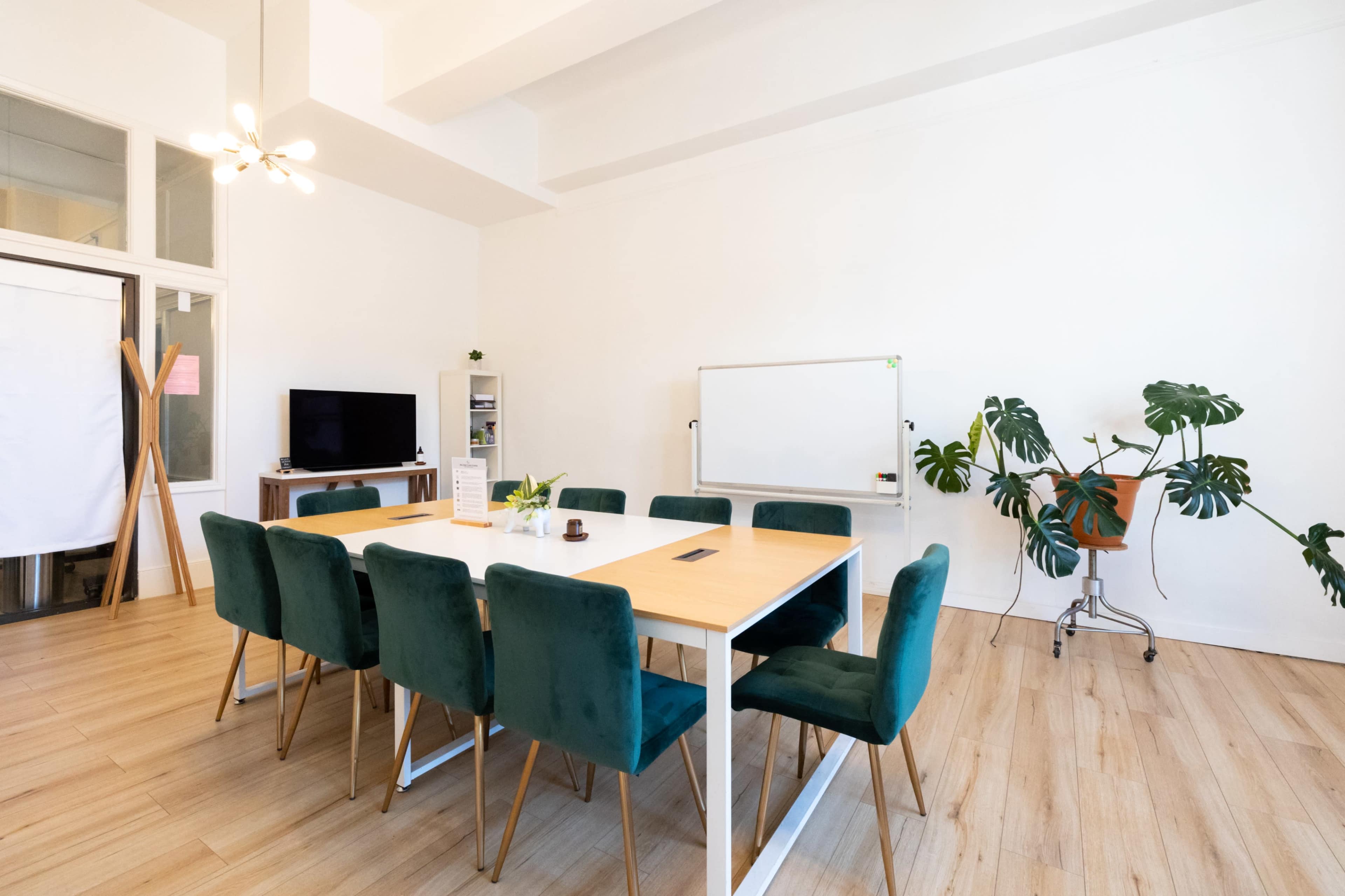 A bright meeting room features a large table surrounded by green velvet chairs, with a whiteboard and large plant in the background.