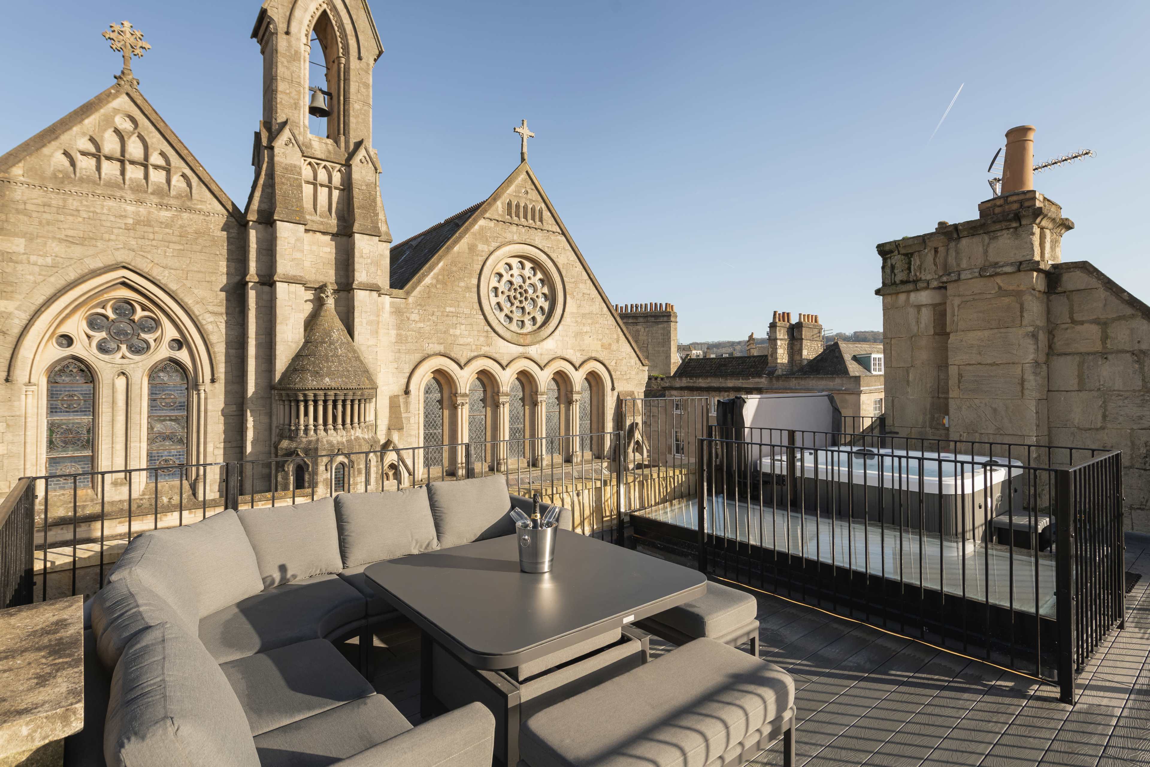 A rooftop terrace features a gray sectional sofa and table overlooking historic stone buildings with decorative architectural details.