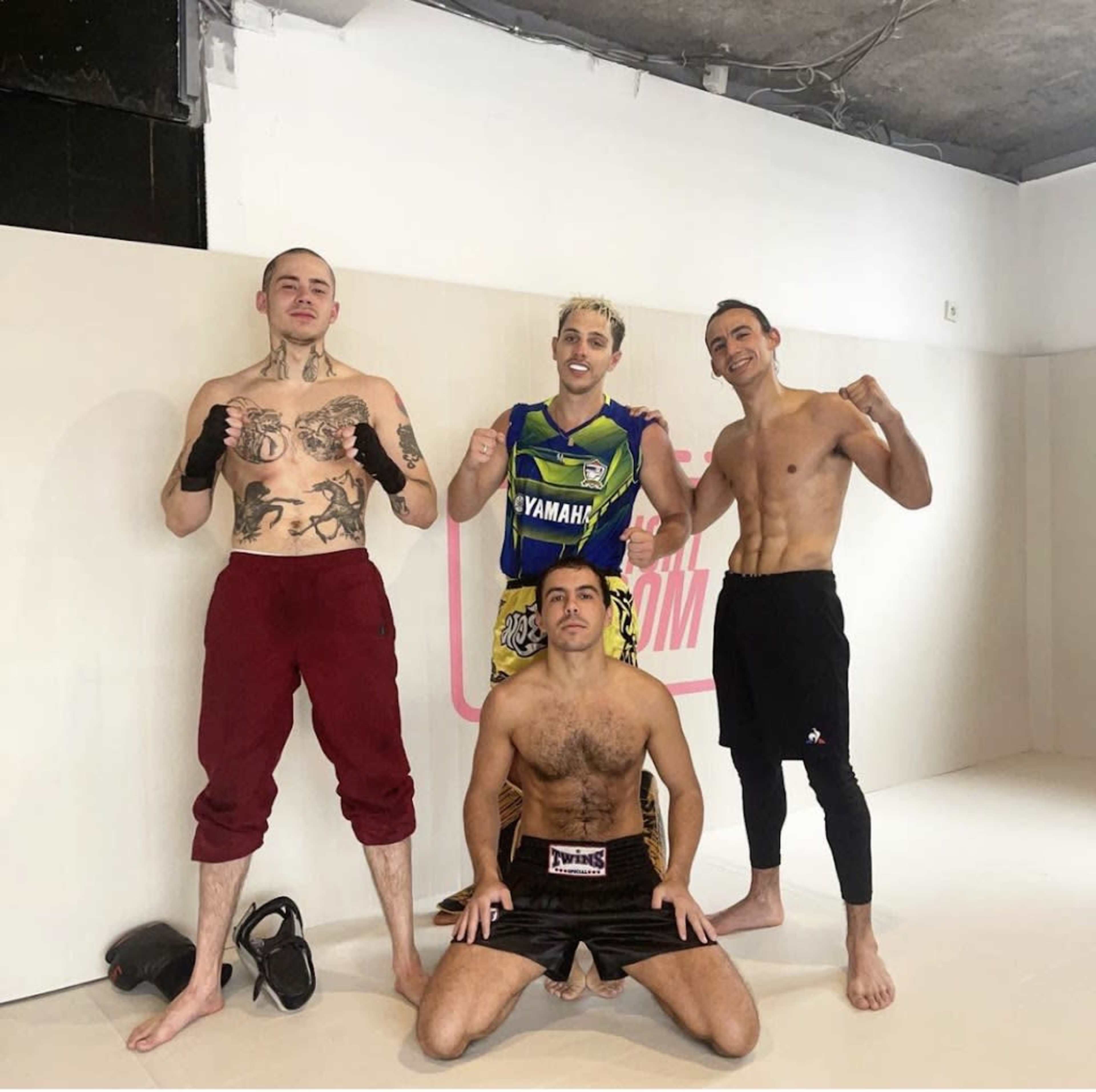 Four shirtless men pose in a training studio, dressed in fight gear and showcasing tattoos.
