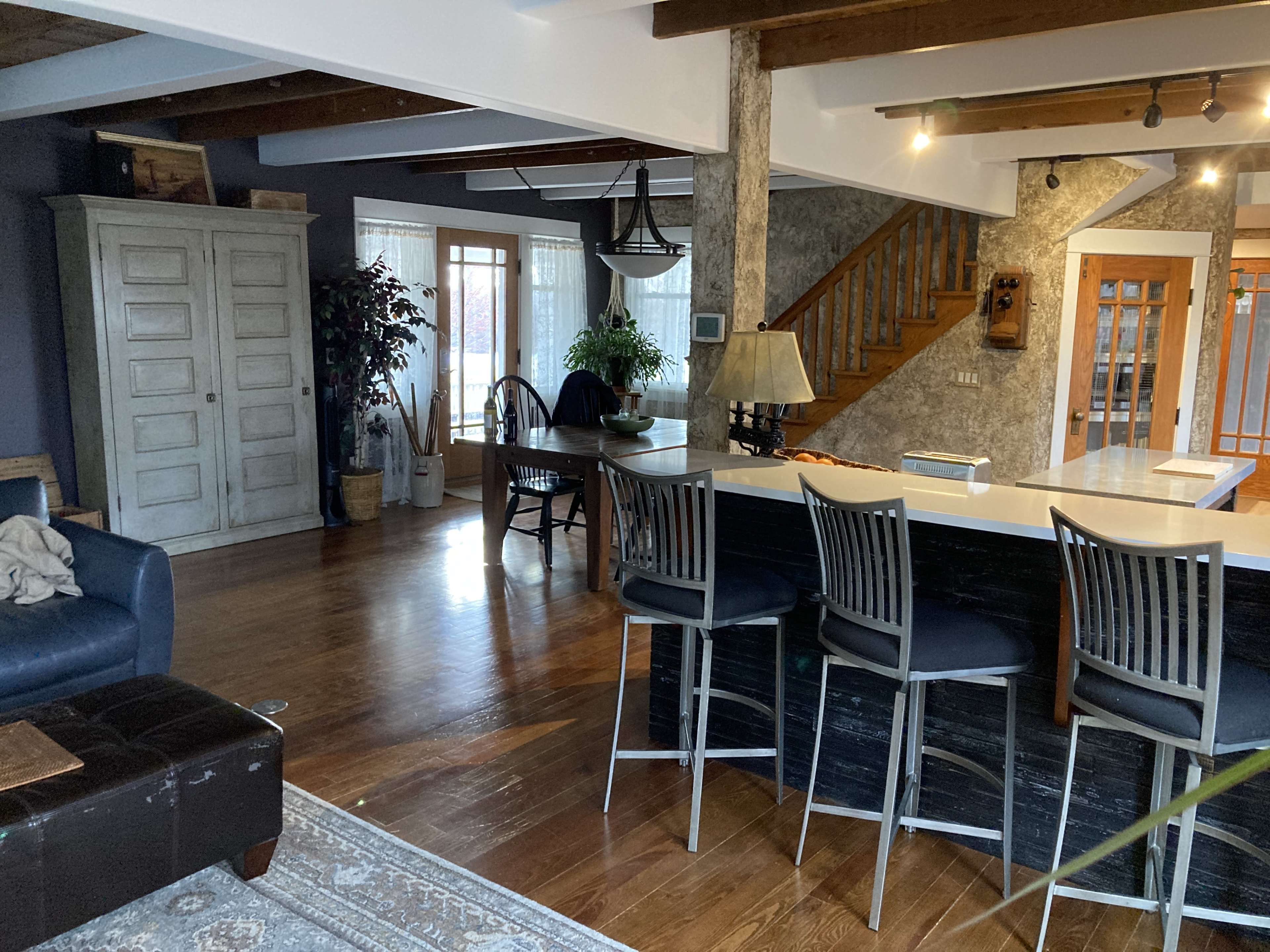 The image shows a spacious living area with a dark sofa, an island bar with stools, a dining table, and a staircase leading to an upper level, illuminated by natural light through large windows.