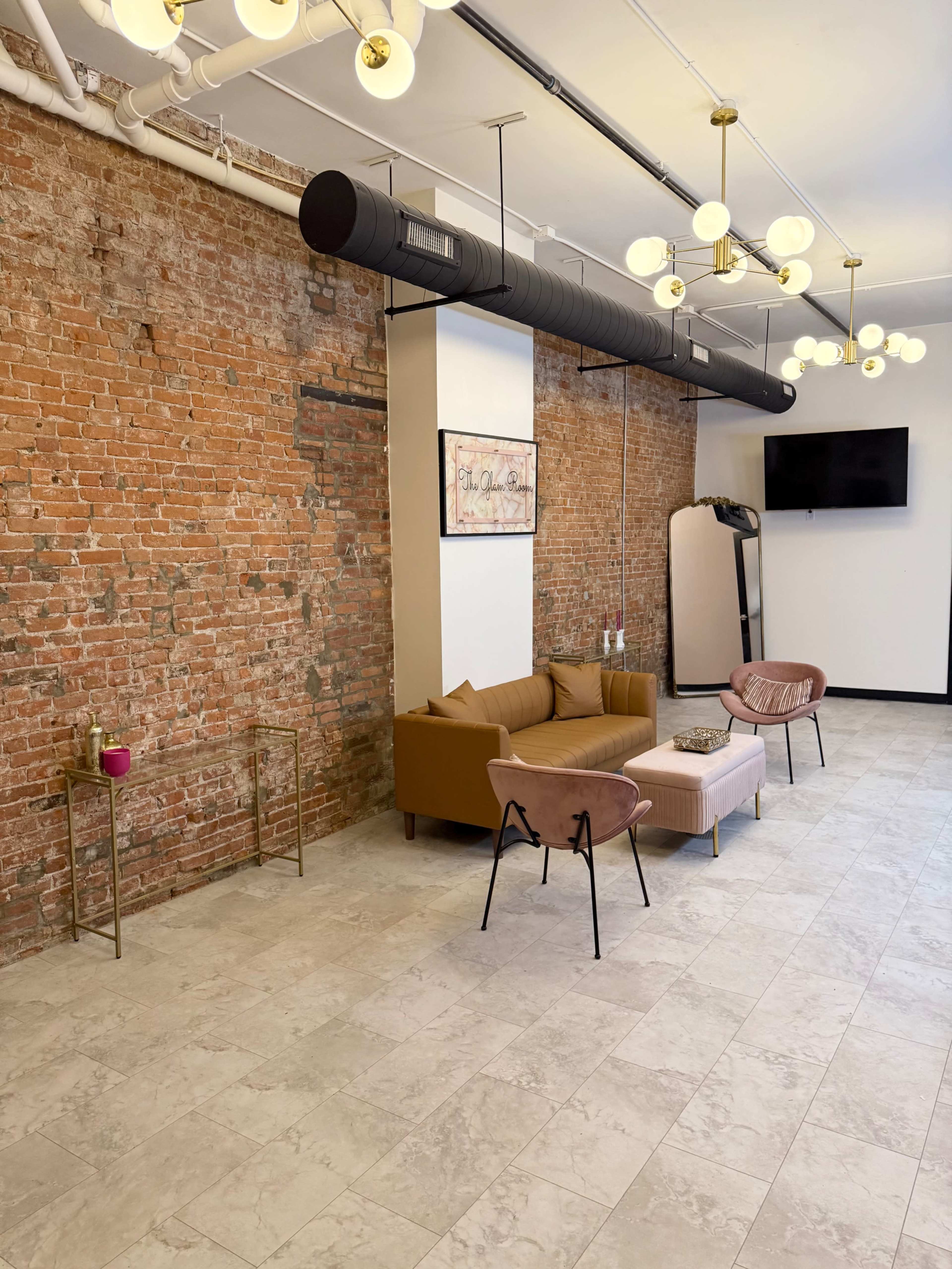 The image shows a modern interior space with exposed brick walls, a brown sofa, two pink chairs, a small table, and a television mounted on the wall.