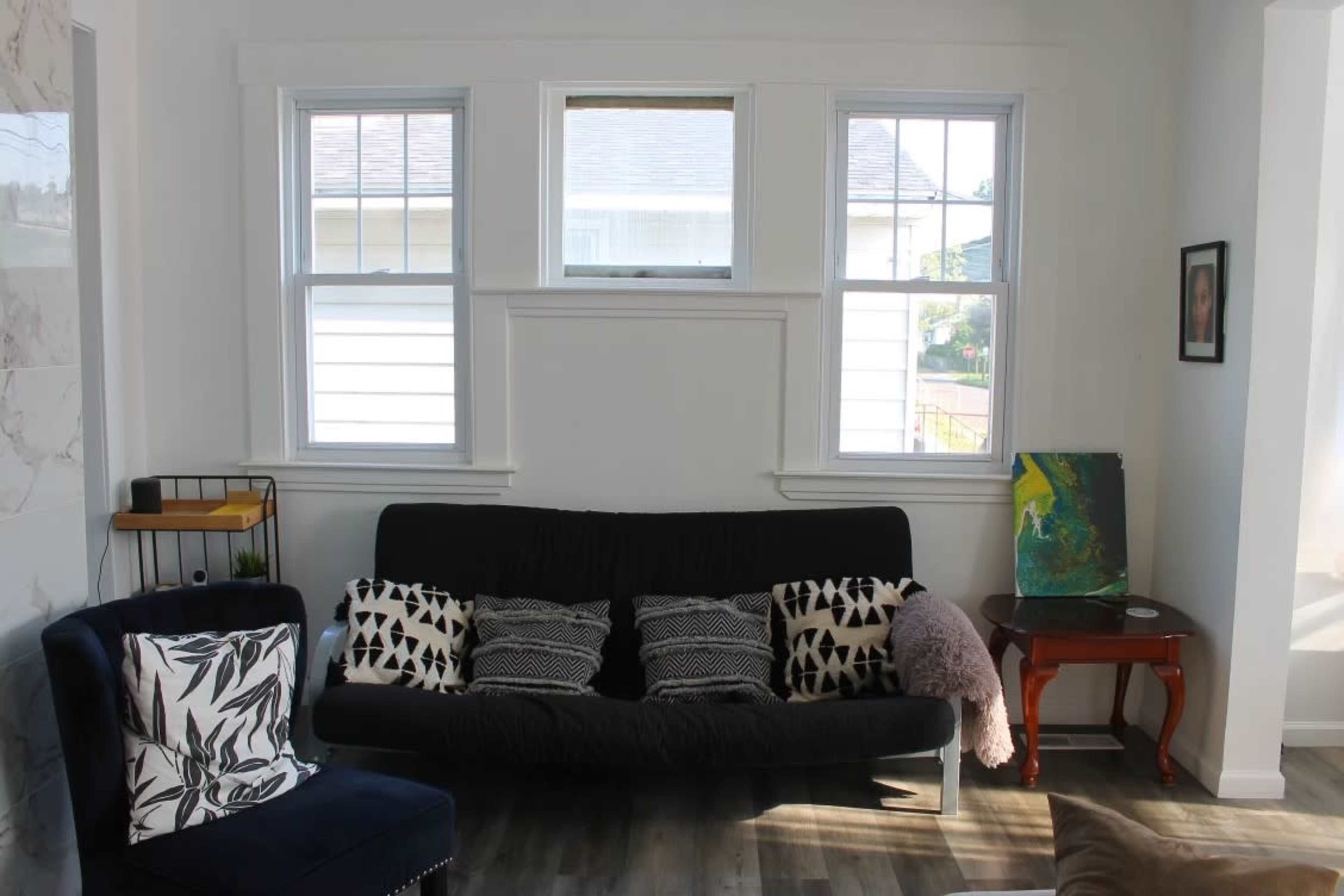 A cozy living room with a black futon, patterned cushions, a small wooden table, and windows providing natural light.