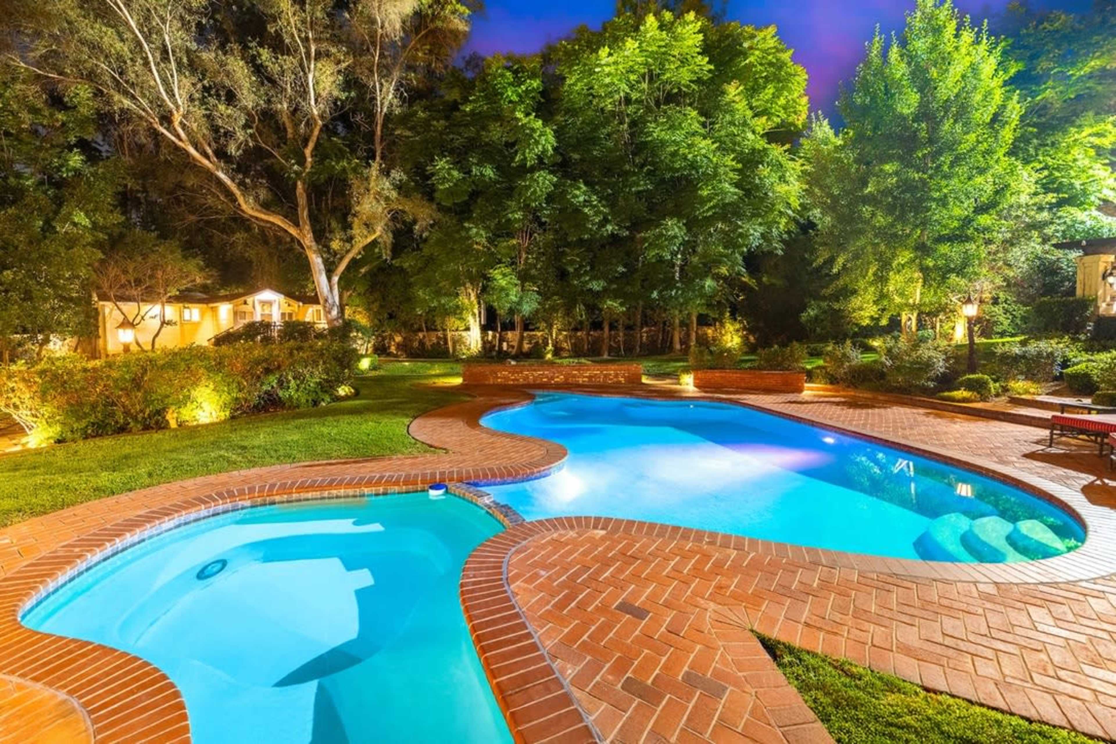 The image shows a brightly lit pool area featuring a curved swimming pool with a spa, surrounded by lush greenery and trees at twilight.