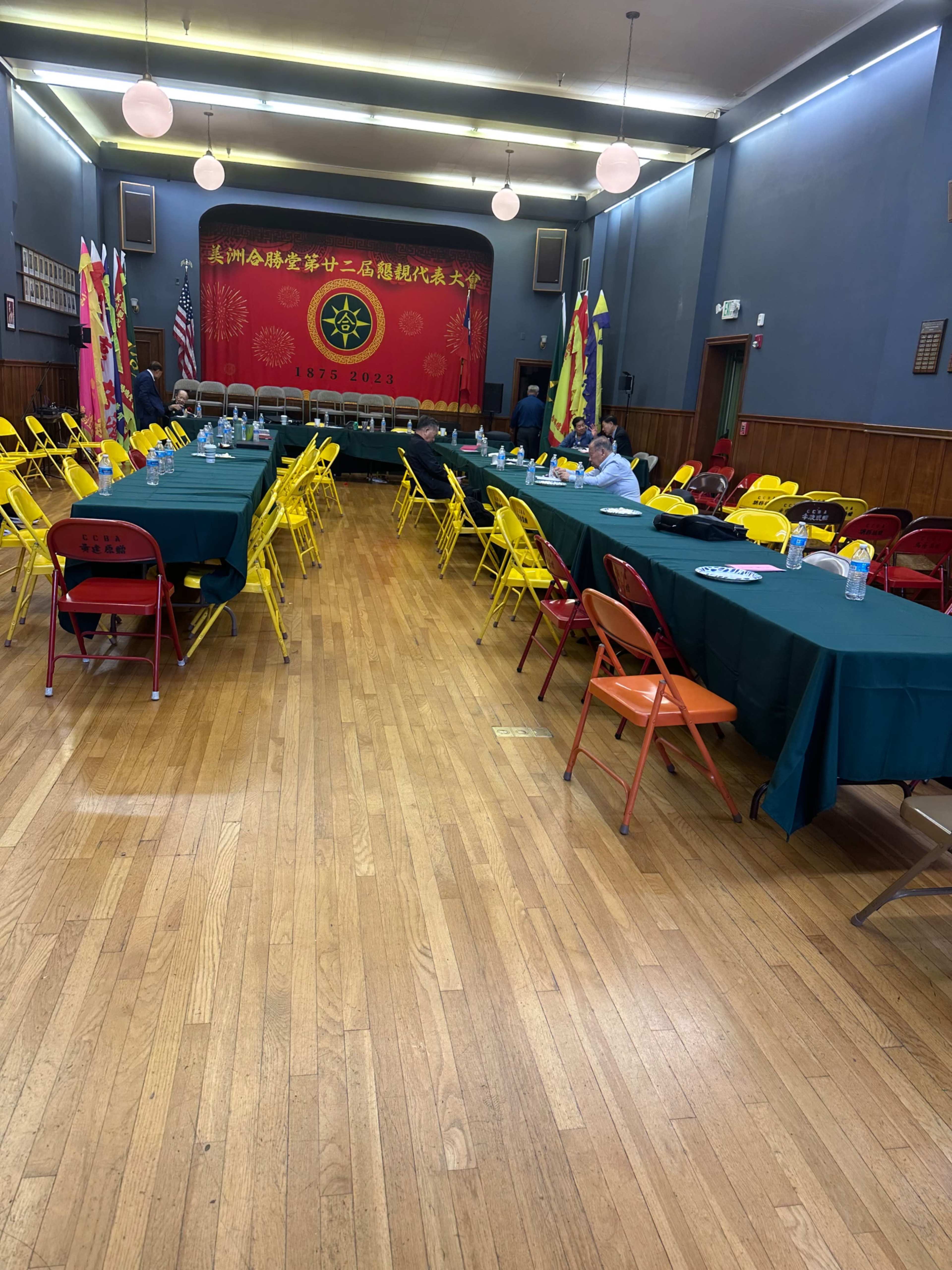 A spacious event hall with yellow and orange chairs arranged around tables, decorated with colorful flags and a large red banner at the front.