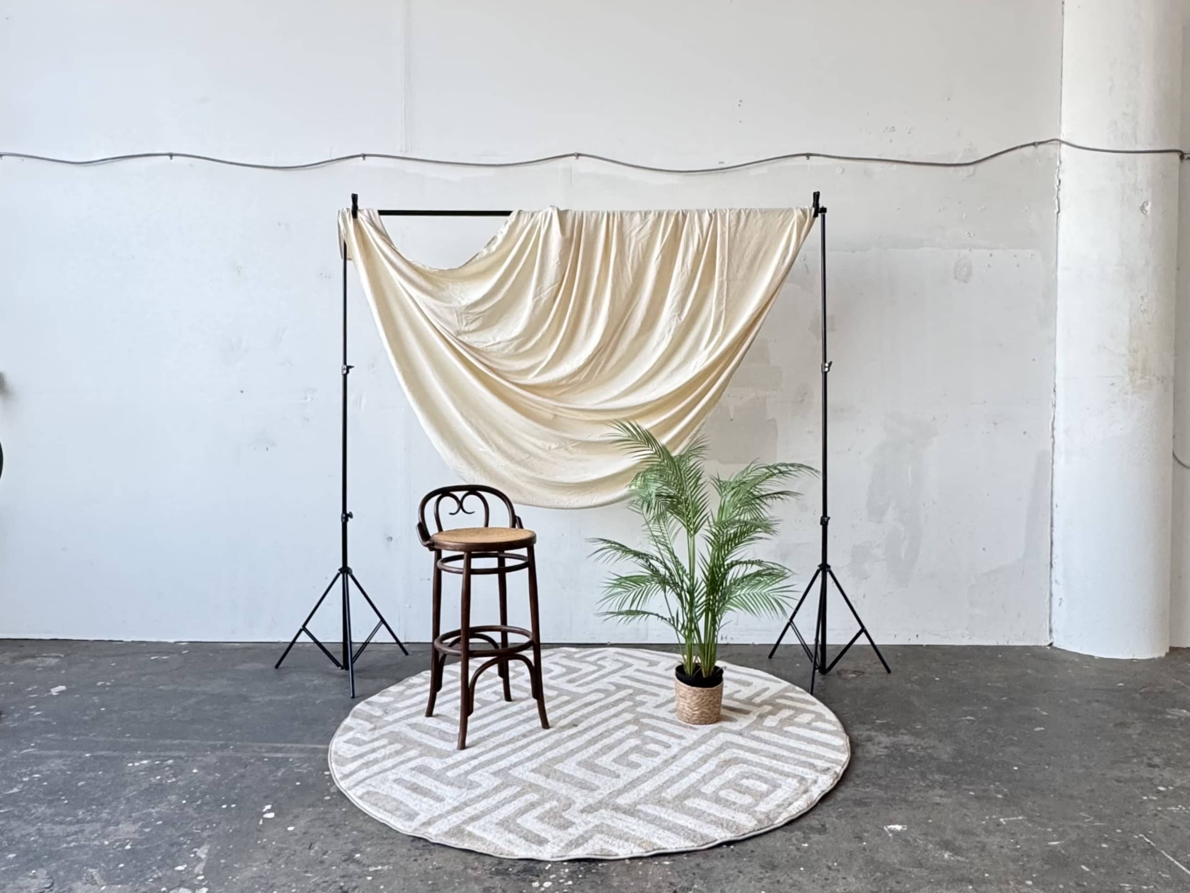 A brown stool is positioned in front of a cream-colored draped backdrop, with a potted plant on the floor and a patterned rug underneath.